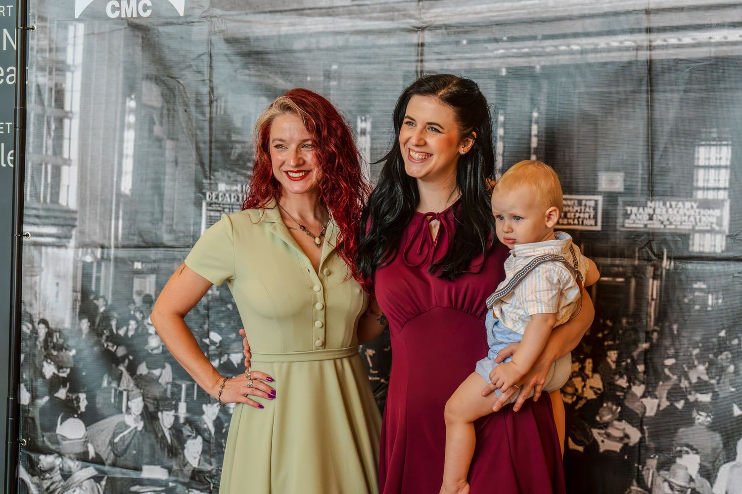 Two women in vintage dresses pose with a baby boy in front of a black and white historical photo backdrop.