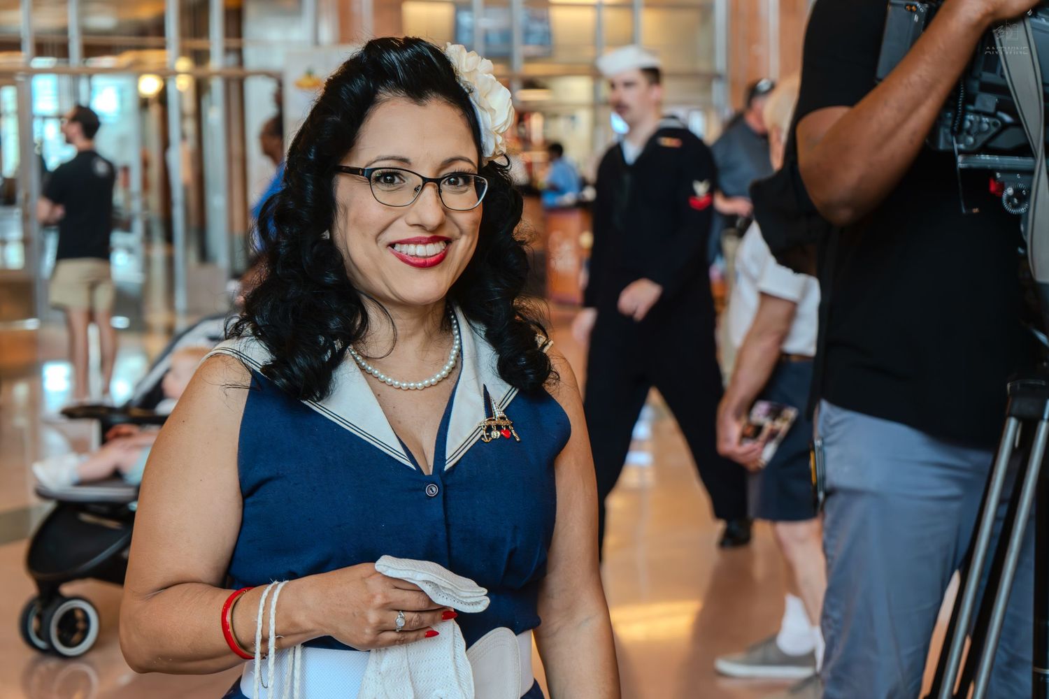 Smiling woman in blue sailor outfit with rainbow pin holding paper at indoor event.