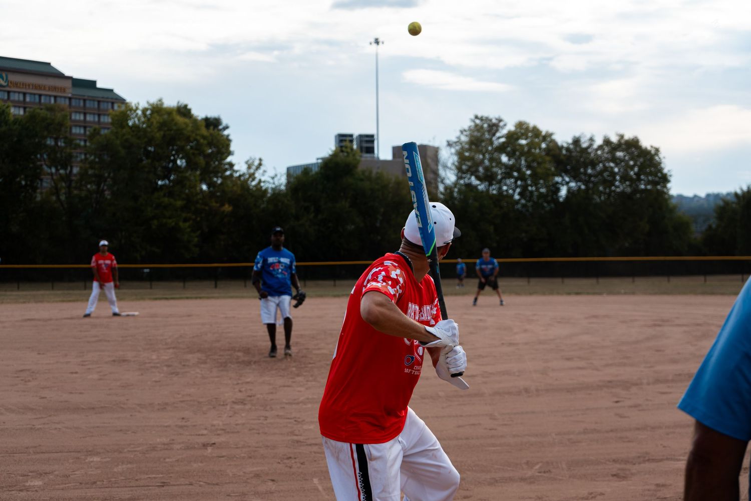 A batter in a red jersey swings at a yellow softball during an outdoor game on a dirt field.