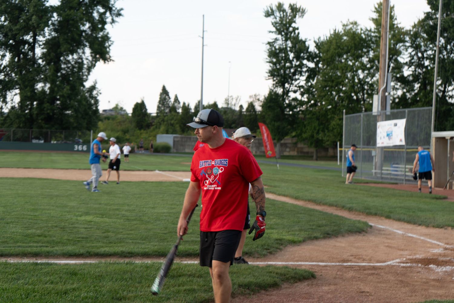 Man in red shirt carries a bat while walking on a baseball diamond during an evening softball game.