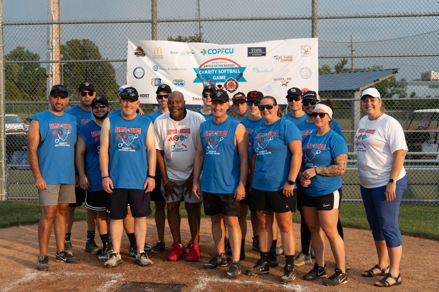 Group of adults in blue team shirts posing on softball field at charity tournament with Comcast sponsor banner.