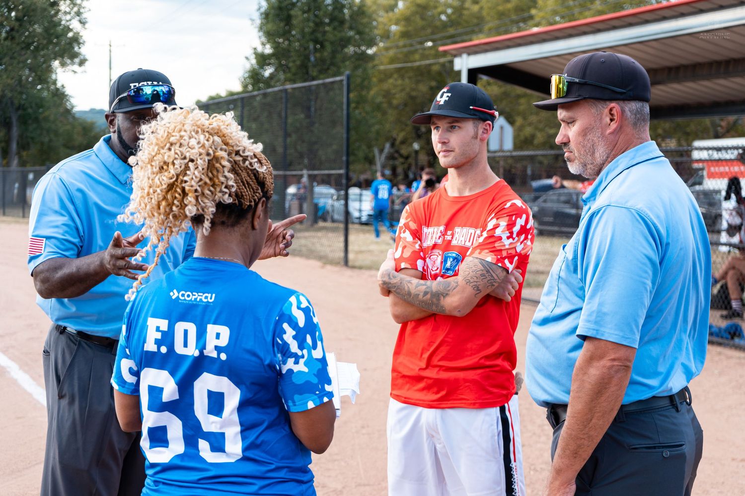 Four people conversing on a softball field, one wearing a blue FOP 69 jersey during an outdoor sports event.