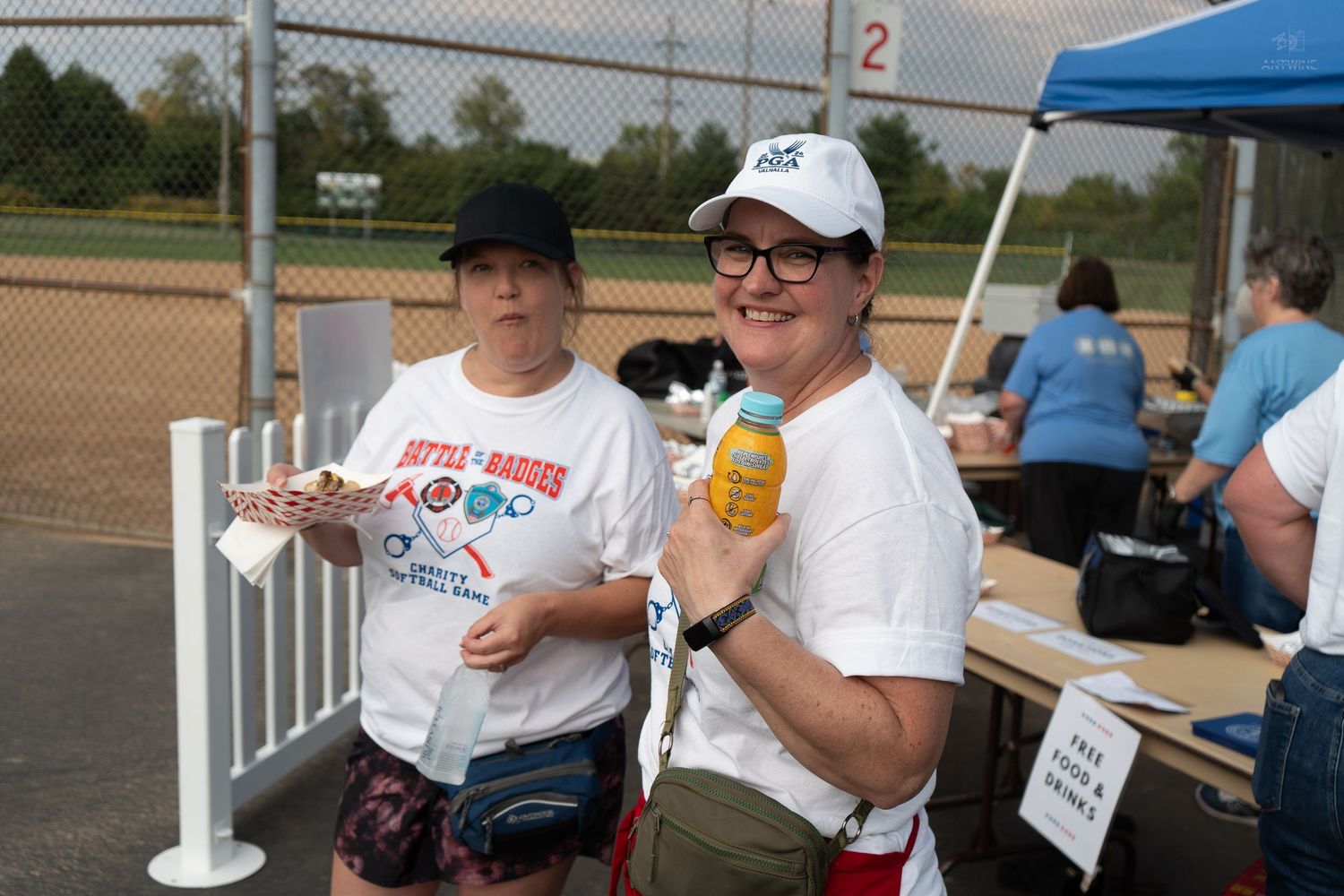 Two women in white caps and shirts smile holding snacks at an outdoor charity softball event.