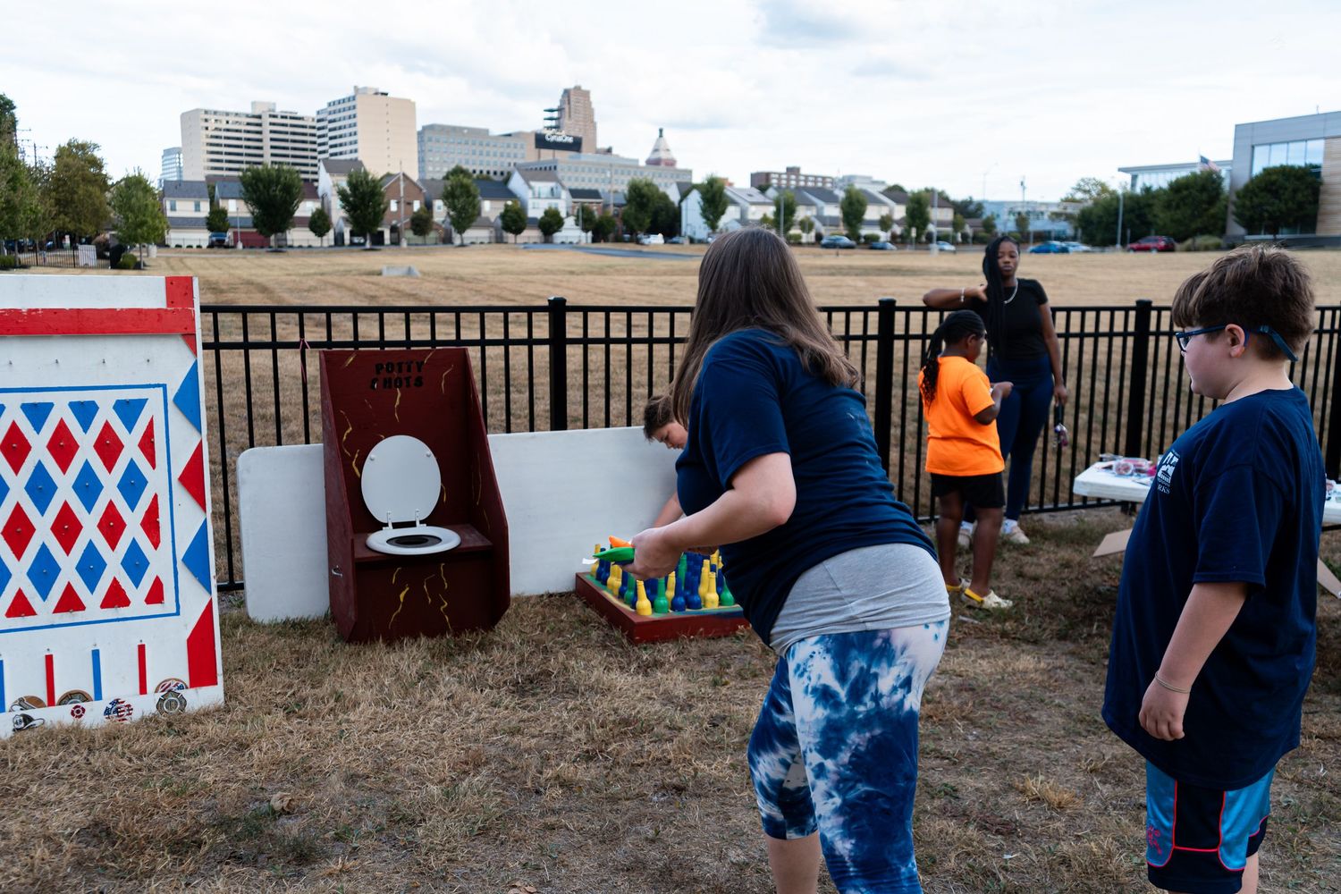 Children play carnival games outdoors at a community festival with city skyline in background.
