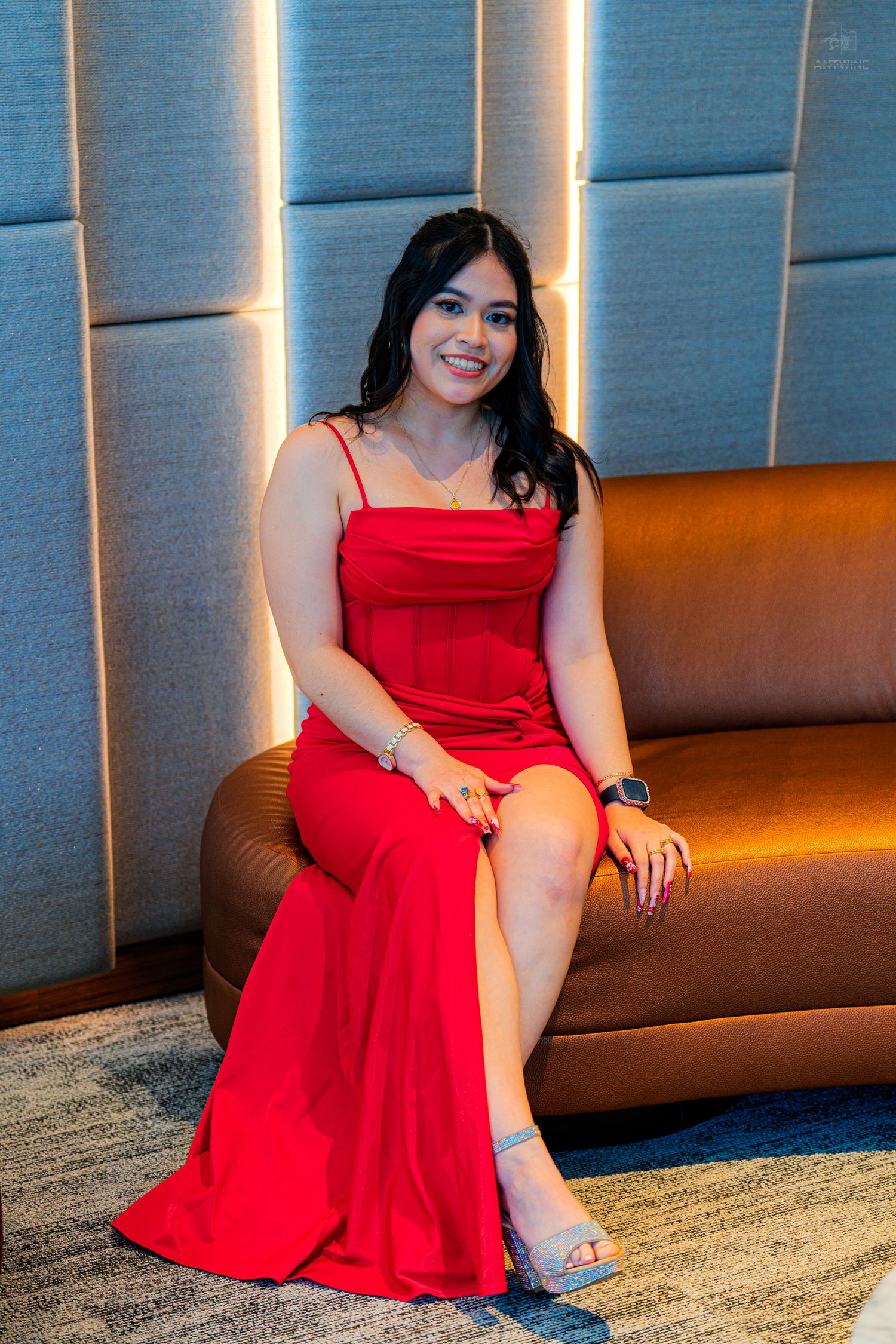 A young woman in a red spaghetti strap gown sits smiling on a brown leather sofa in an elegant room.