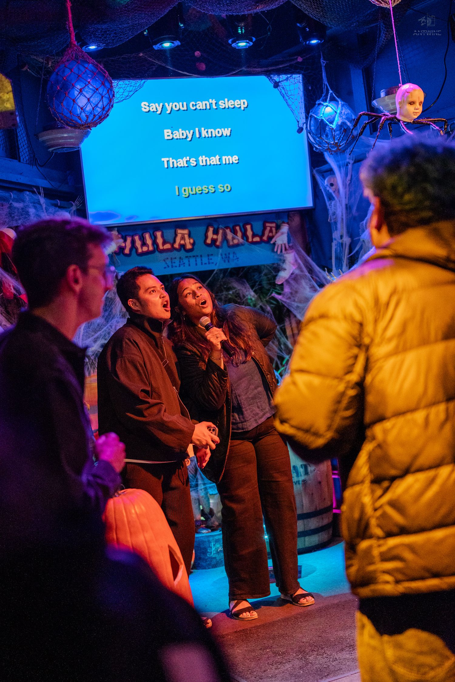 Two people singing karaoke duet at Hula Hula bar with spooky Halloween decorations in Seattle.