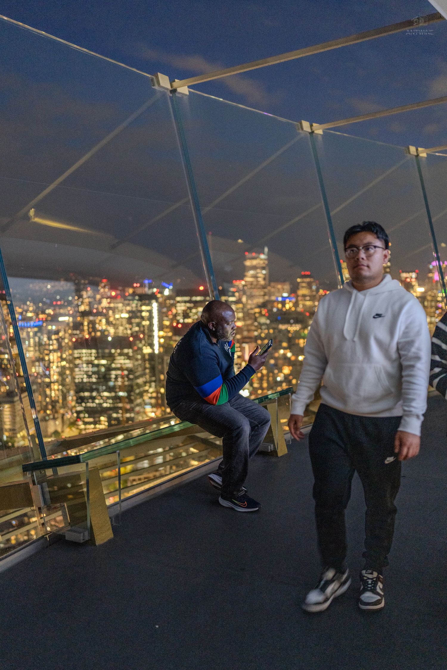 Two men posing on observation deck with colorful city skyline at night.
