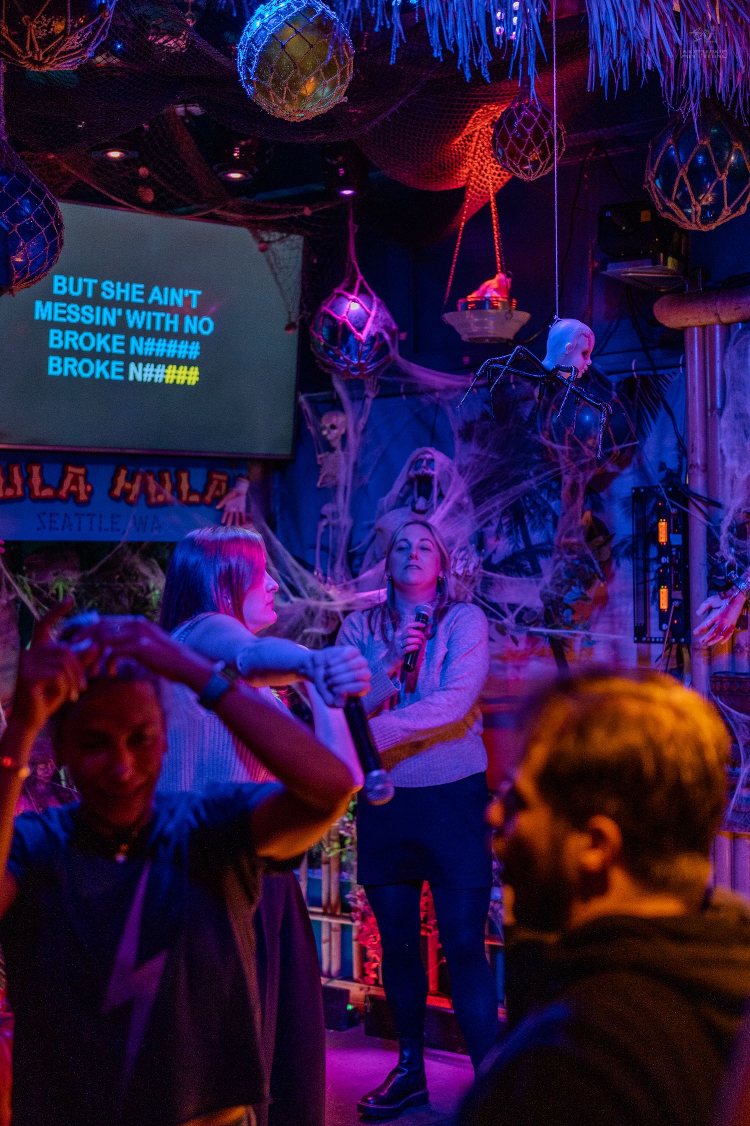 Woman singing karaoke at Hula Hula bar in Seattle with Halloween decorations and neon lights.
