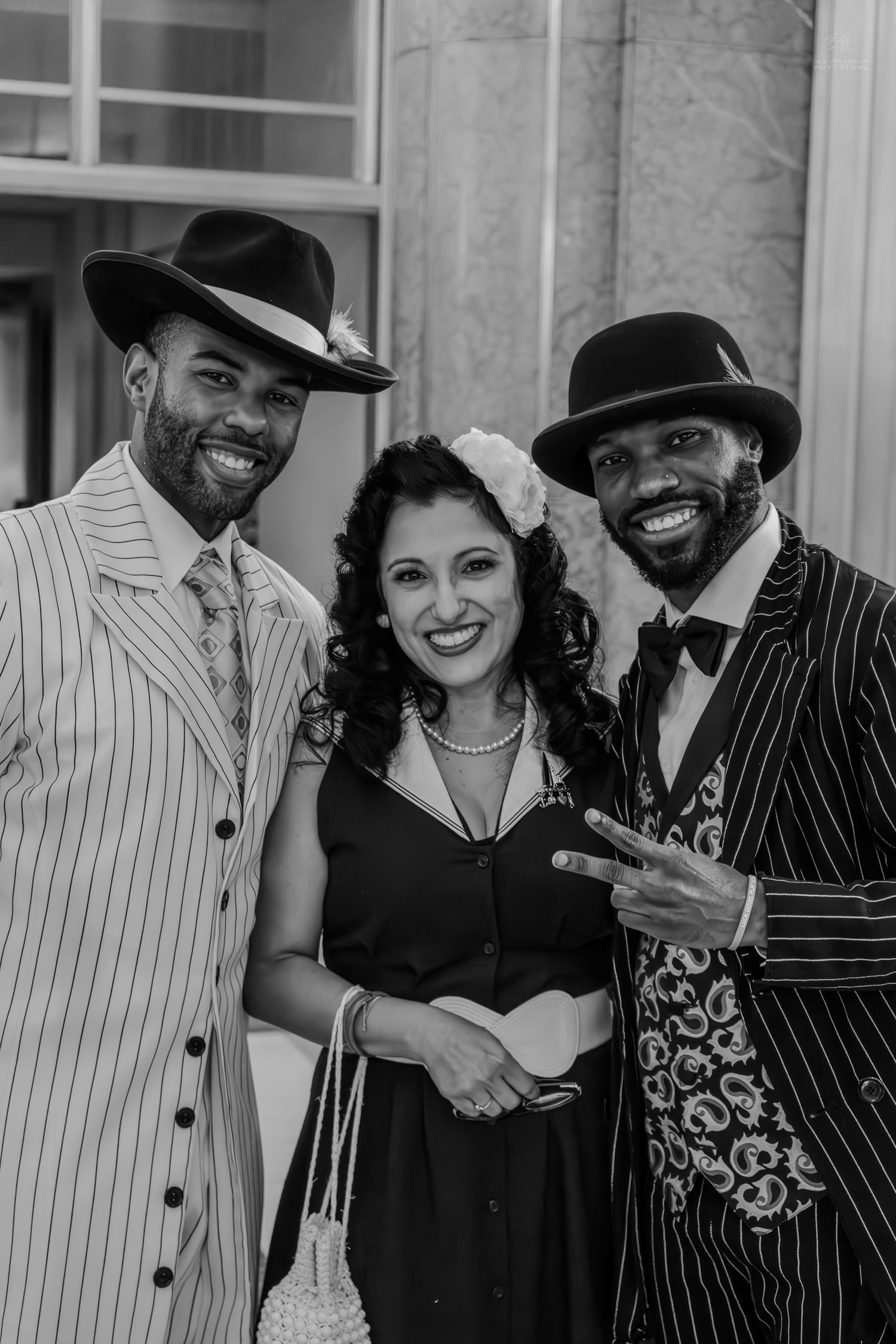 Three stylish people in vintage pinstripe suits and a black dress pose together smiling at a retro-themed event.