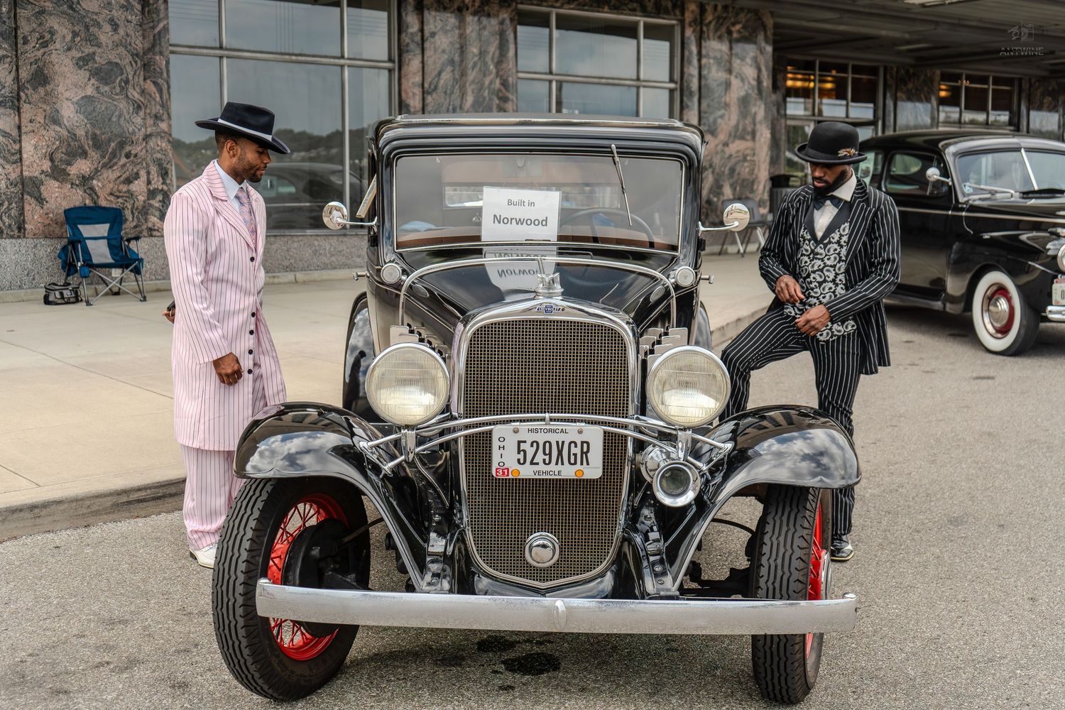 Two men in zoot suits pose with a vintage black classic car at an outdoor car show event.