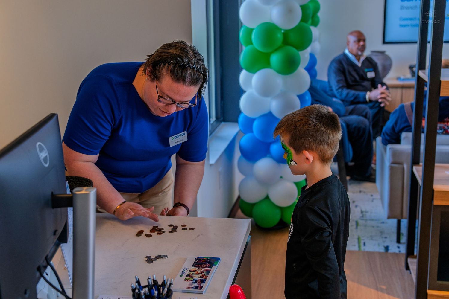 A staff member in a blue shirt assists a child at a activity table during a balloon-decorated celebration event.