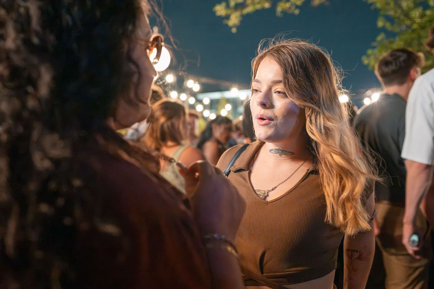 Two women talking at an outdoor evening event with string lights in the background.