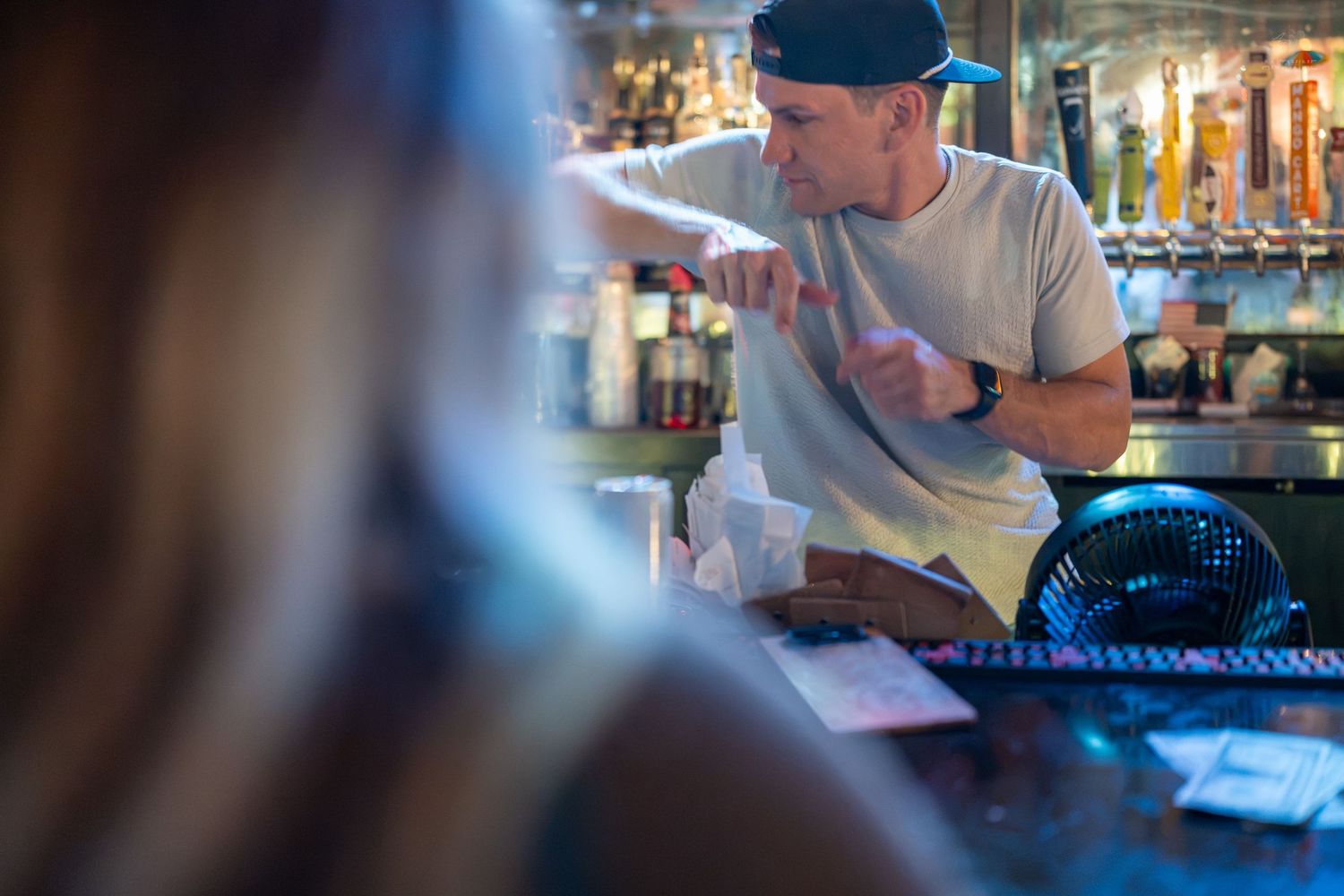 Bartender in gray shirt and cap takes an order from a customer at a busy bar counter.