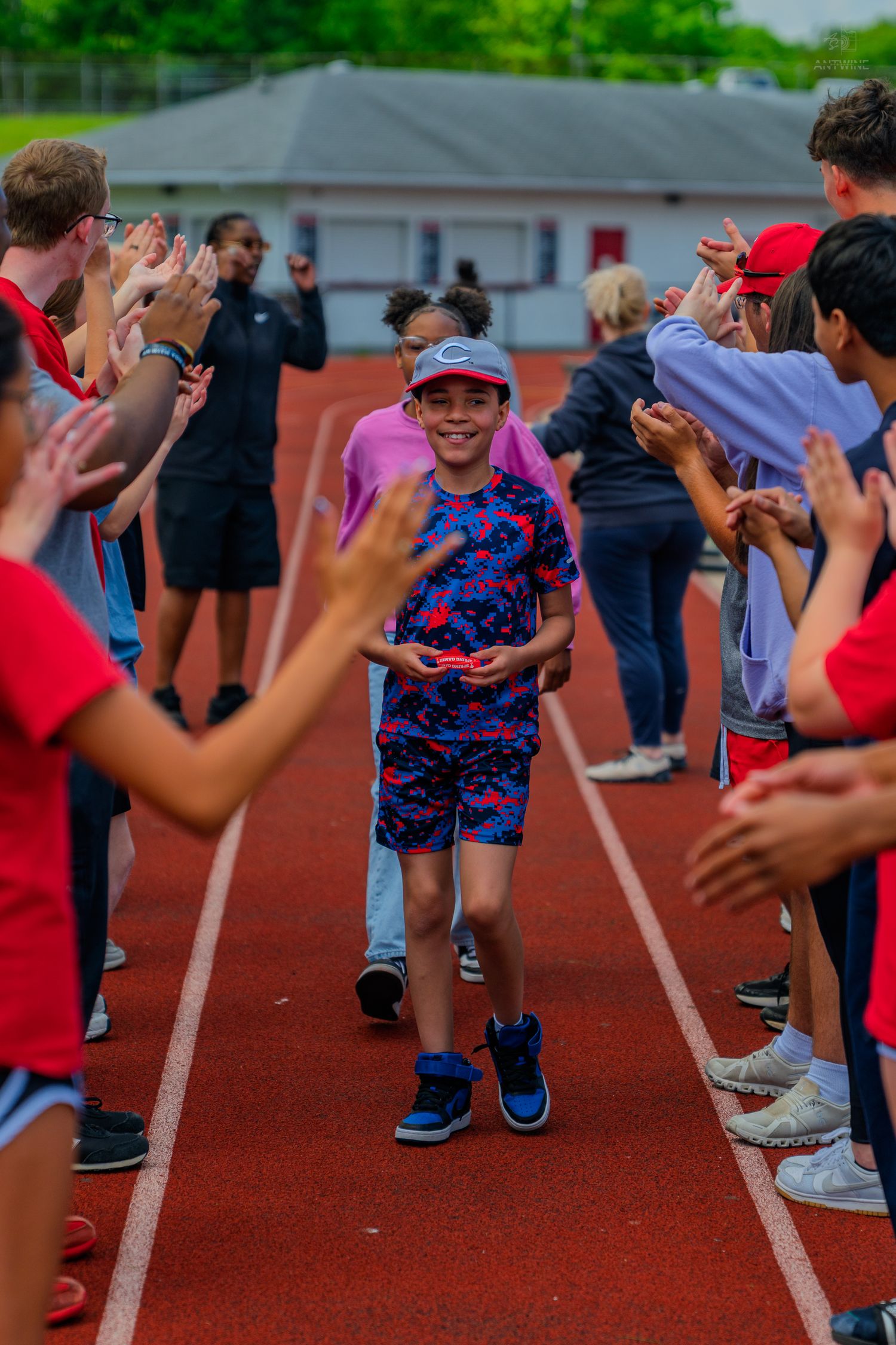 Smiling child walks through a celebratory high-five tunnel on a red track surrounded by cheering peers.