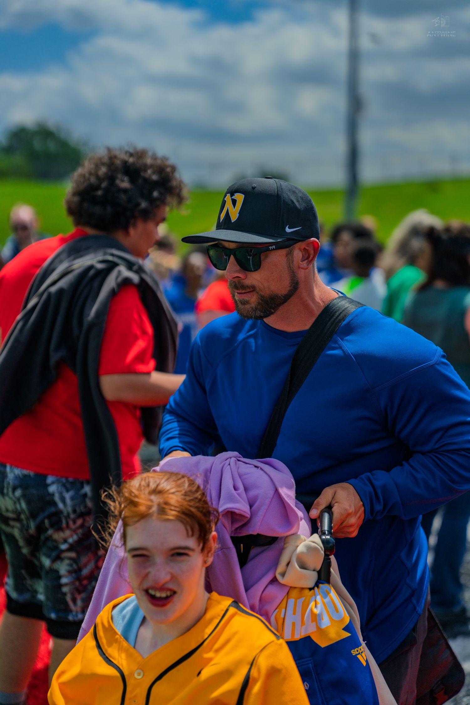 A man in a blue shirt and cap pushes a child in a wheelchair at an outdoor event.