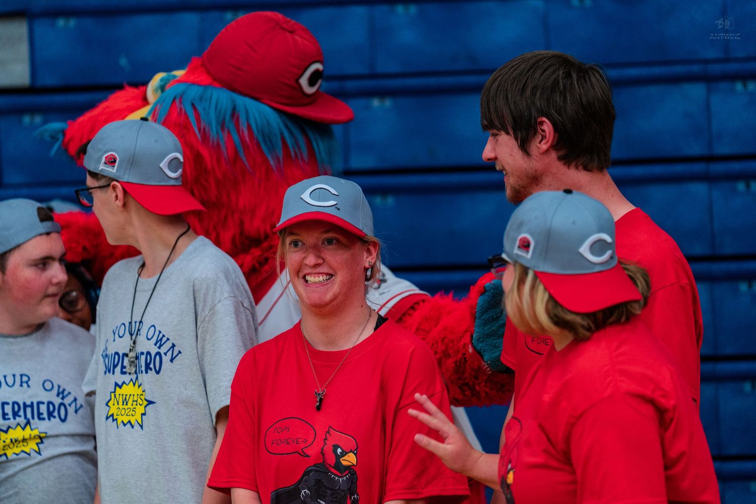 Group of smiling participants pose with Cincinnati Reds mascot Mr. Red wearing team caps at an event.