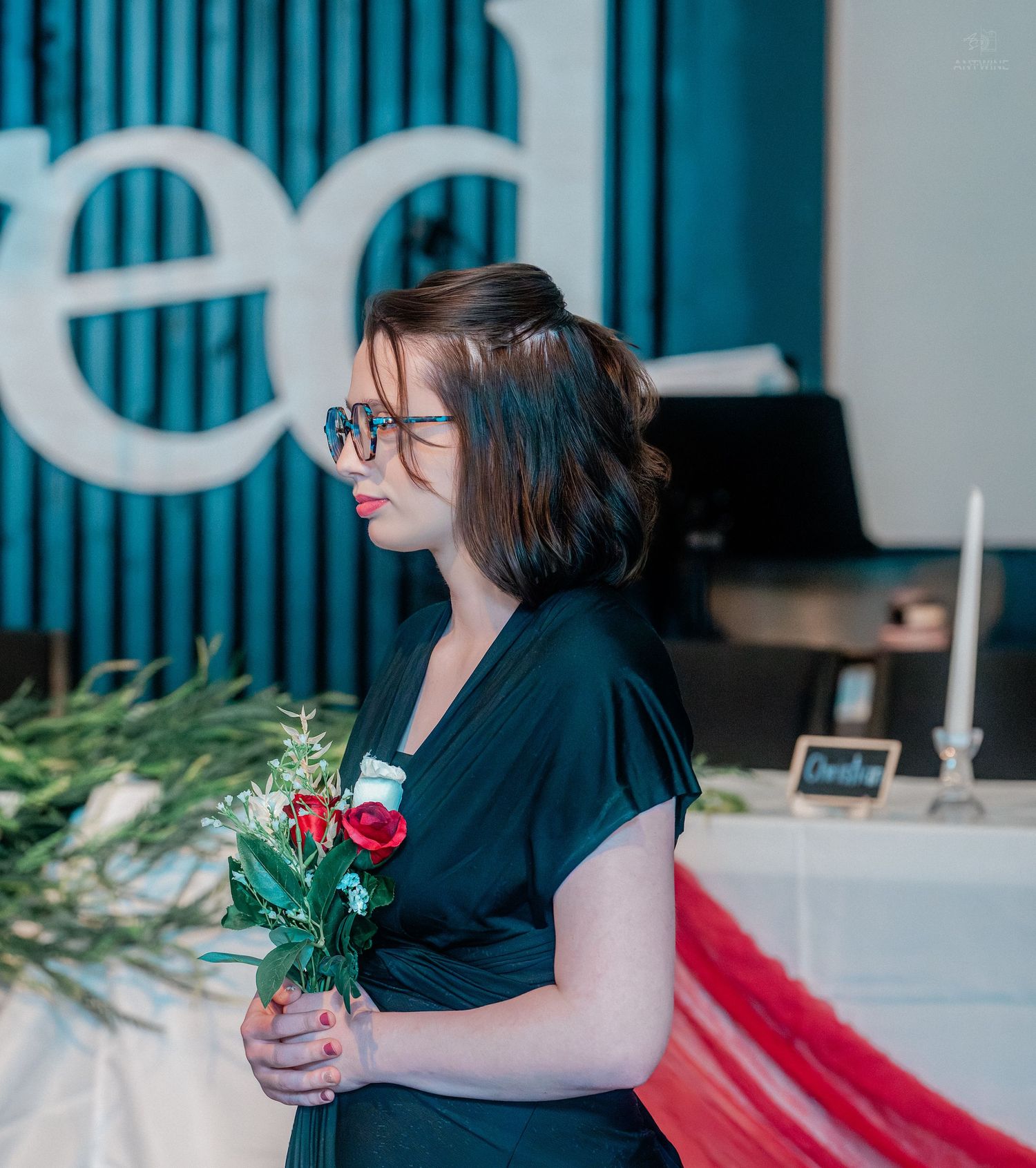 Bridesmaid in black dress holds red rose bouquet while standing near wedding reception decor.