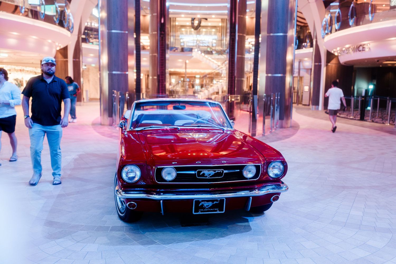 A classic red Ford Mustang convertible on display inside a modern cruise ship atrium.