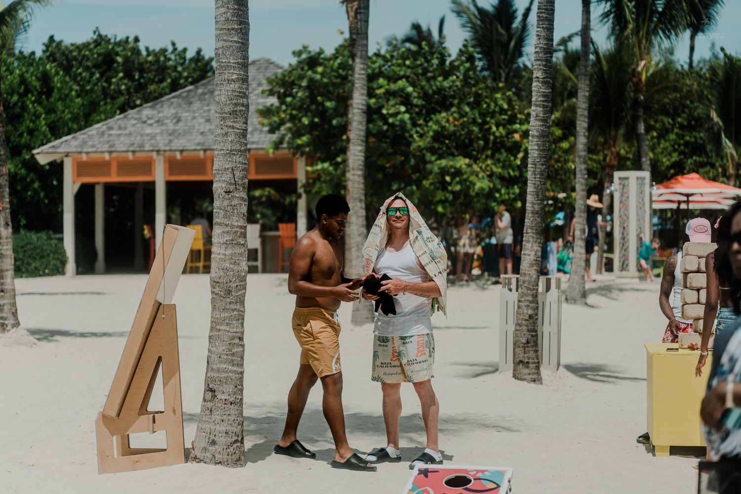 Two people examining camera equipment near a cornhole game on a tropical beach resort with palm trees.