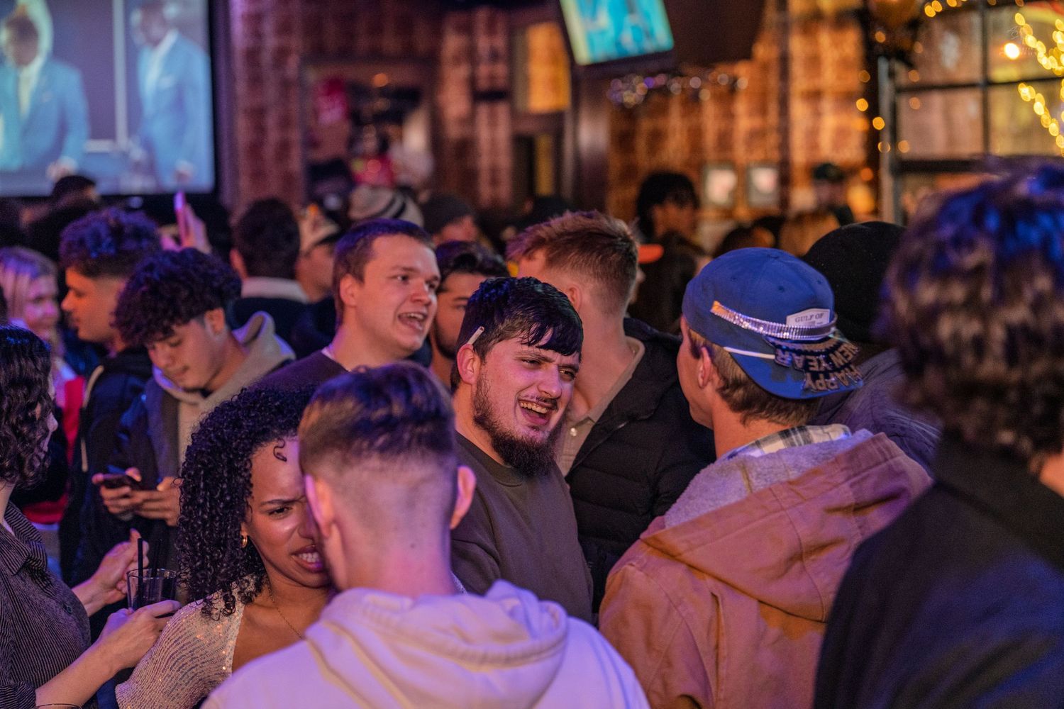 Group of young adults talking and socializing in a lively crowded bar with TV screens.