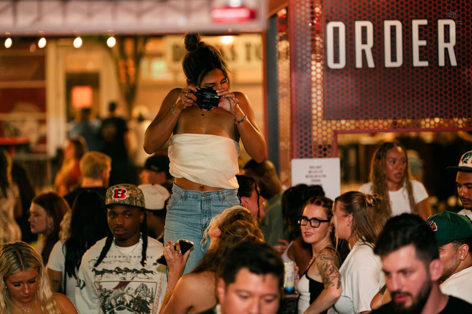 Woman photographing crowd at busy indoor food market event with ORDER sign visible.