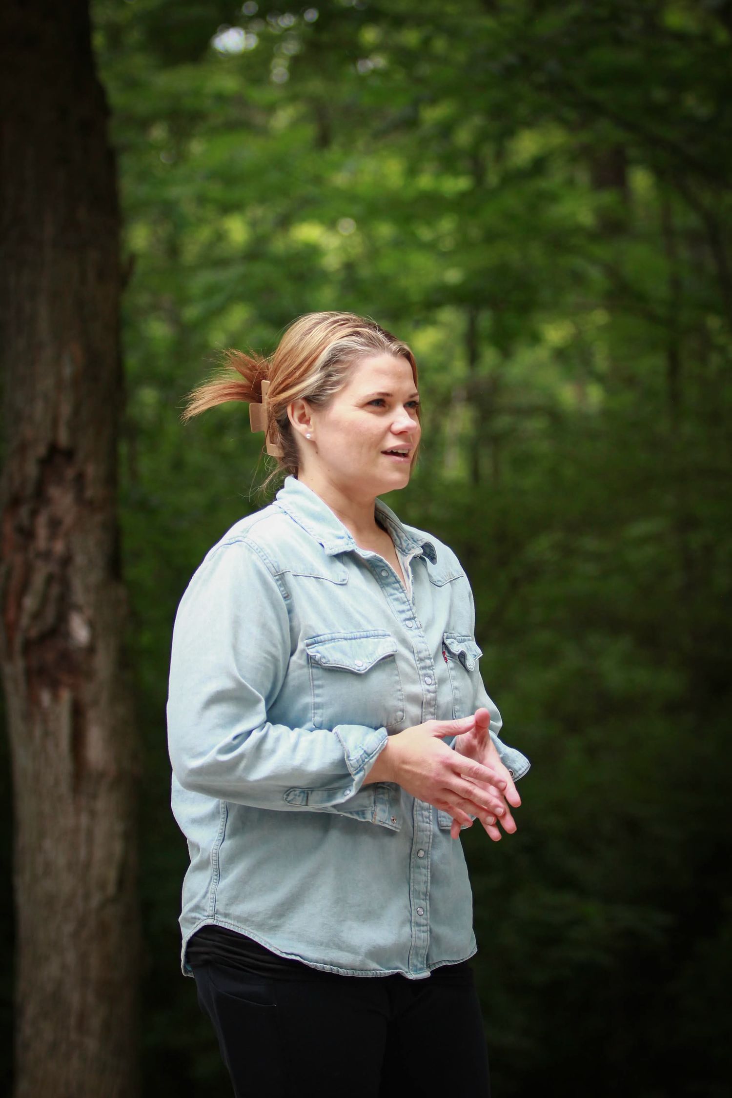 Woman in denim shirt speaking outdoors in a lush green forest setting.