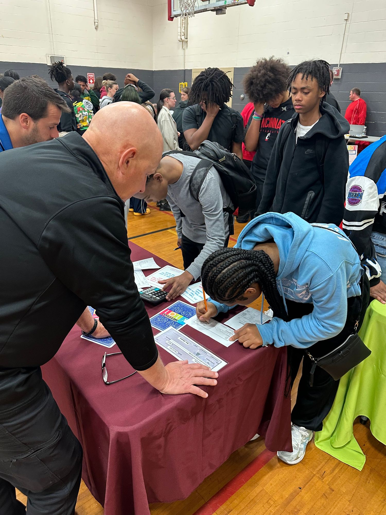 Students sign up at a table during a school career fair event in a gymnasium with a bald man assisting.