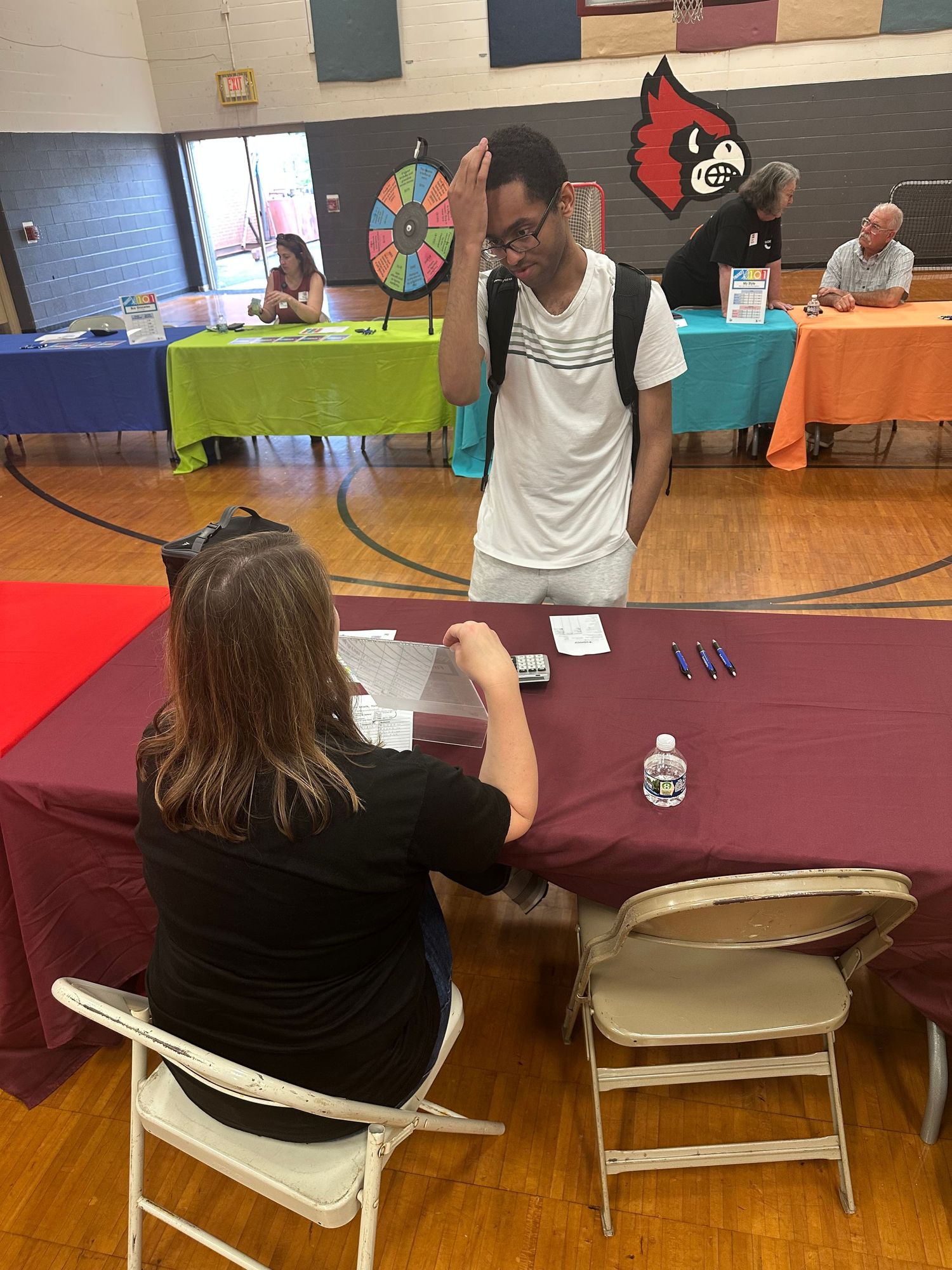 Students interact at a school career fair in a gymnasium with colorful tables and a cardinal mascot logo.