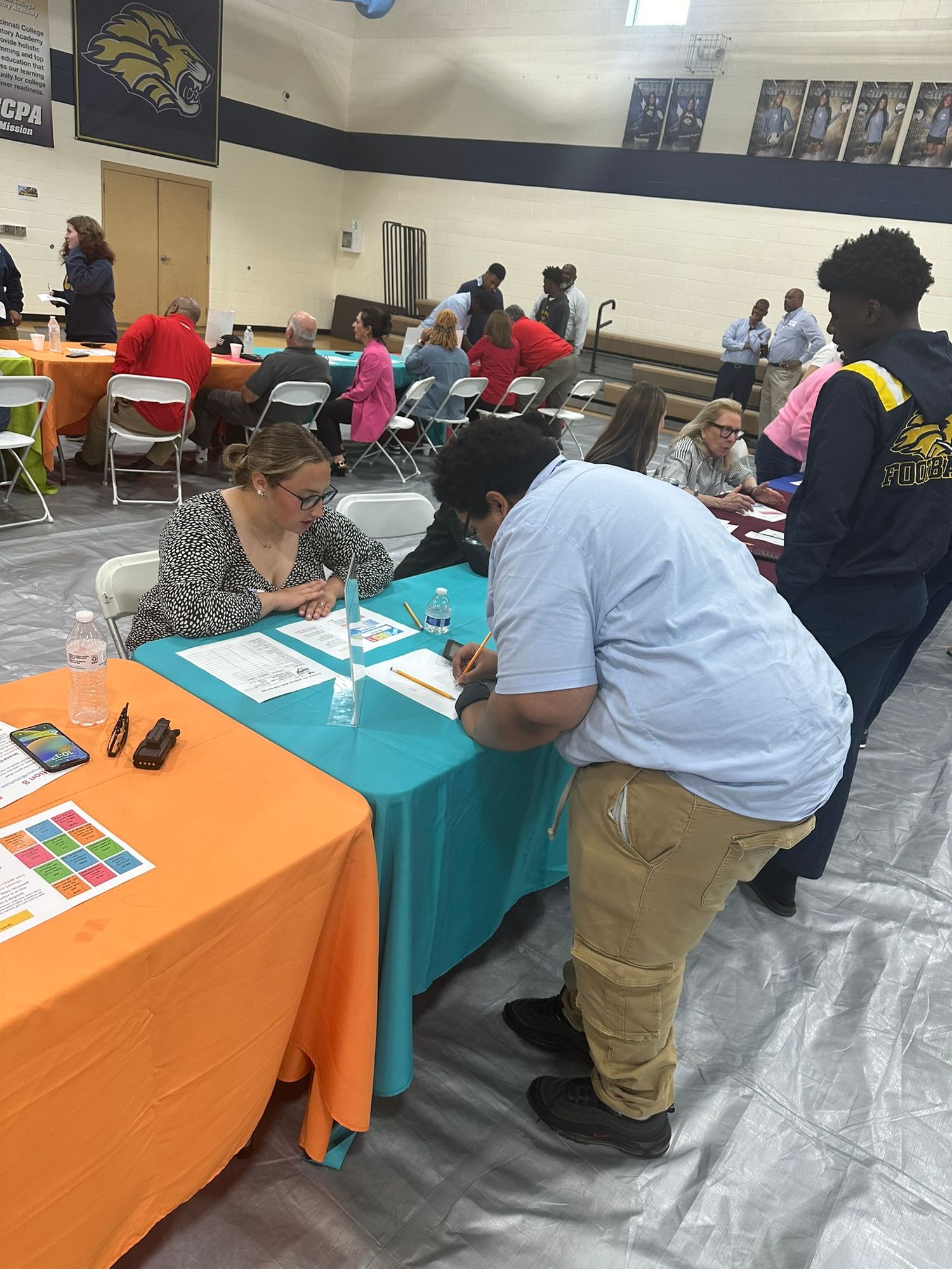 Students and recruiters interact at indoor career fair with colorful tables in school gymnasium.