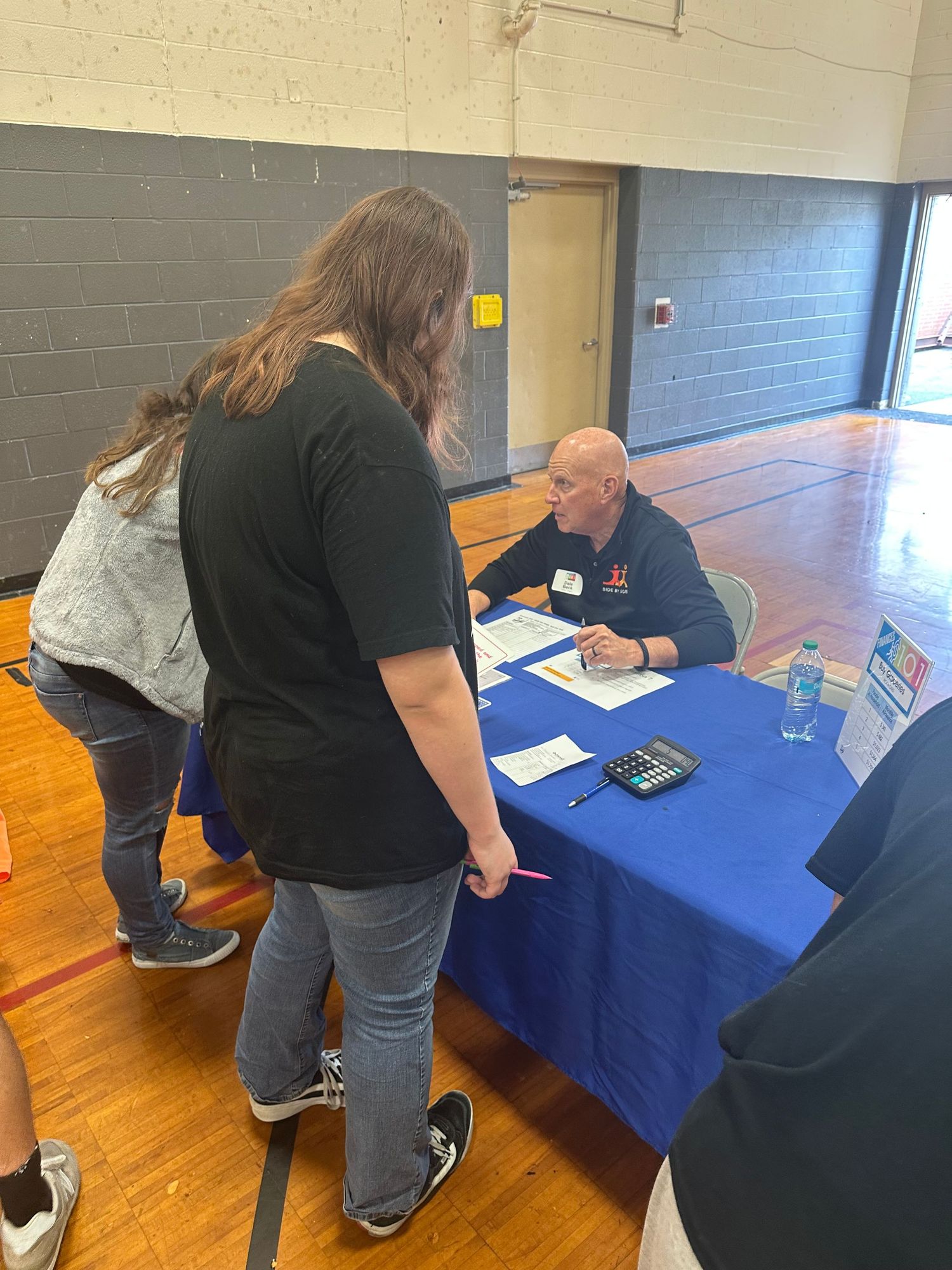 People speaking with a recruiter at a blue table during a job fair in a gymnasium.