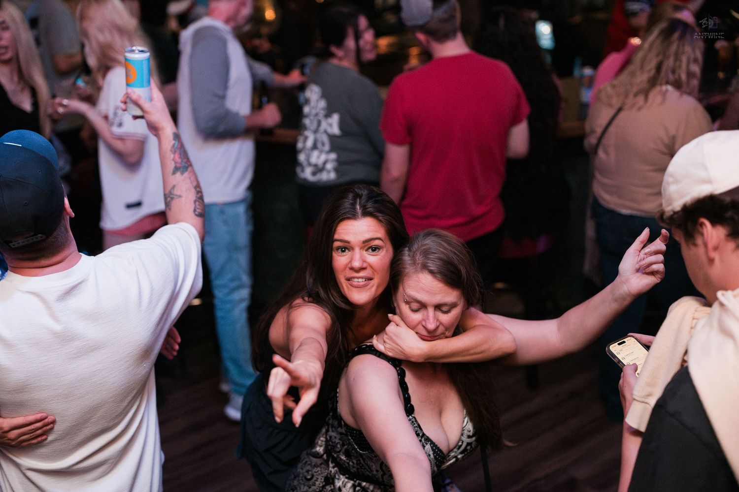 Two women posing and smiling together at a lively crowded bar or nightclub event.