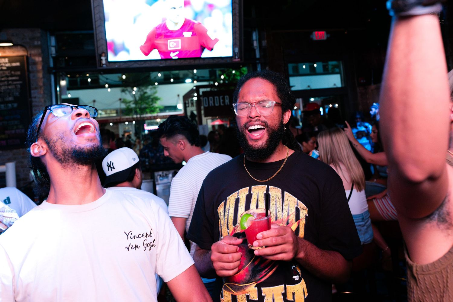 Two men laughing and holding cocktails in front of a large screen at a lively nightlife venue.