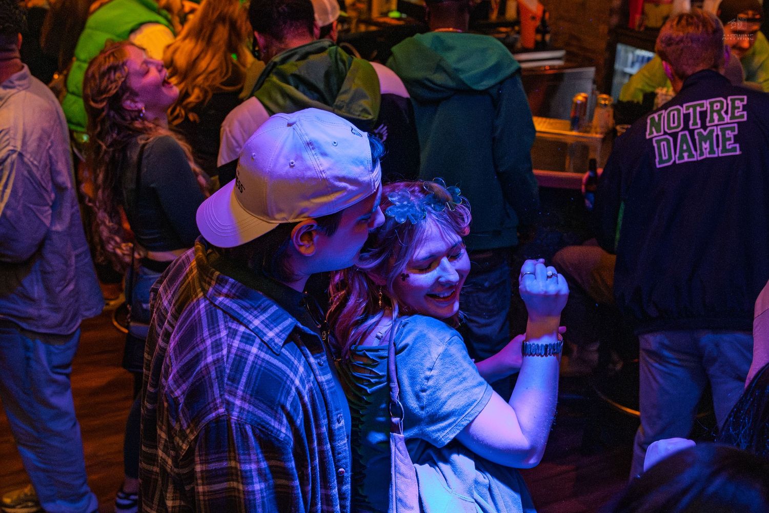Couple dancing together at a crowded bar party with Notre Dame shirt visible in background.