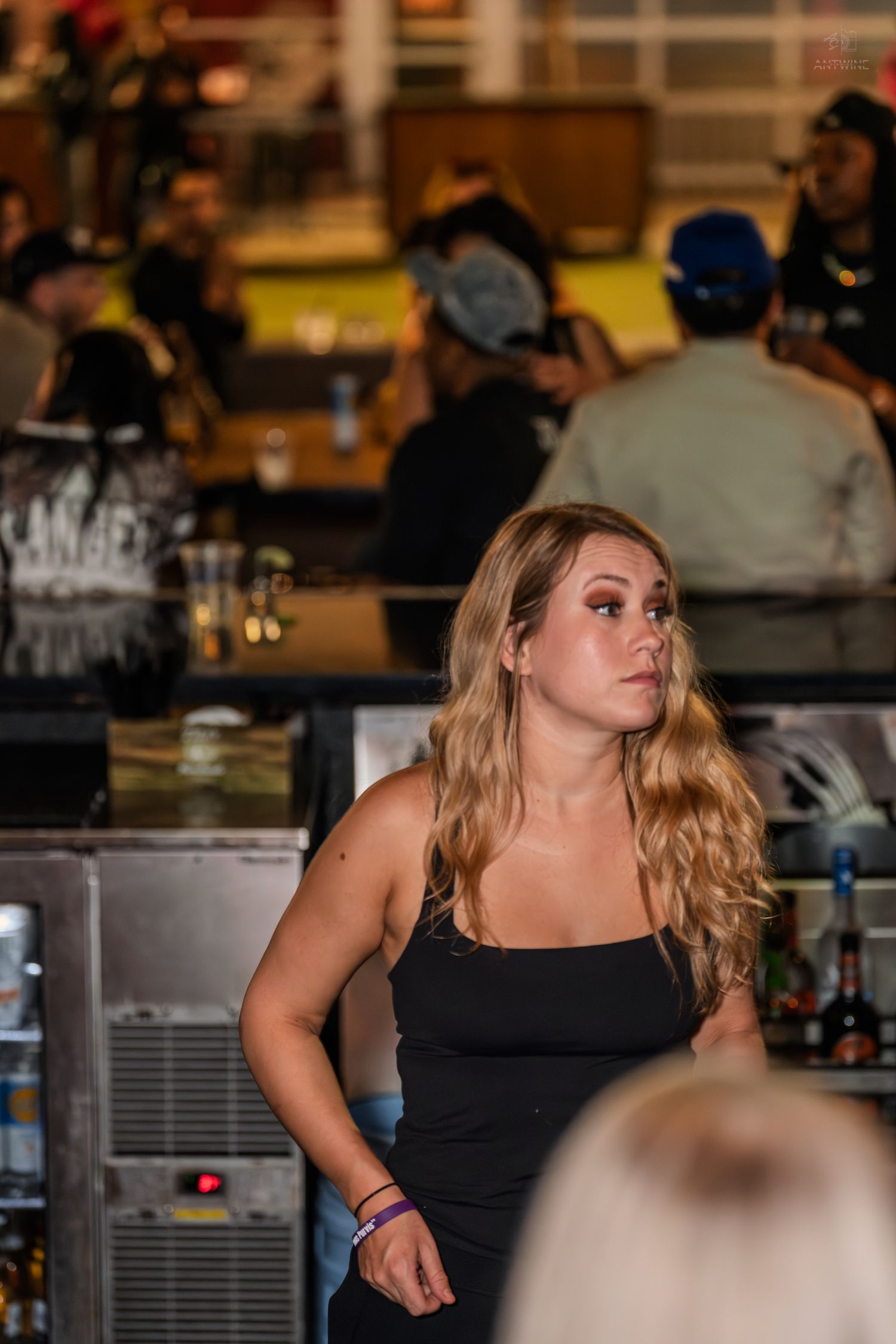 Blonde bartender in black dress standing attentively behind a busy bar.