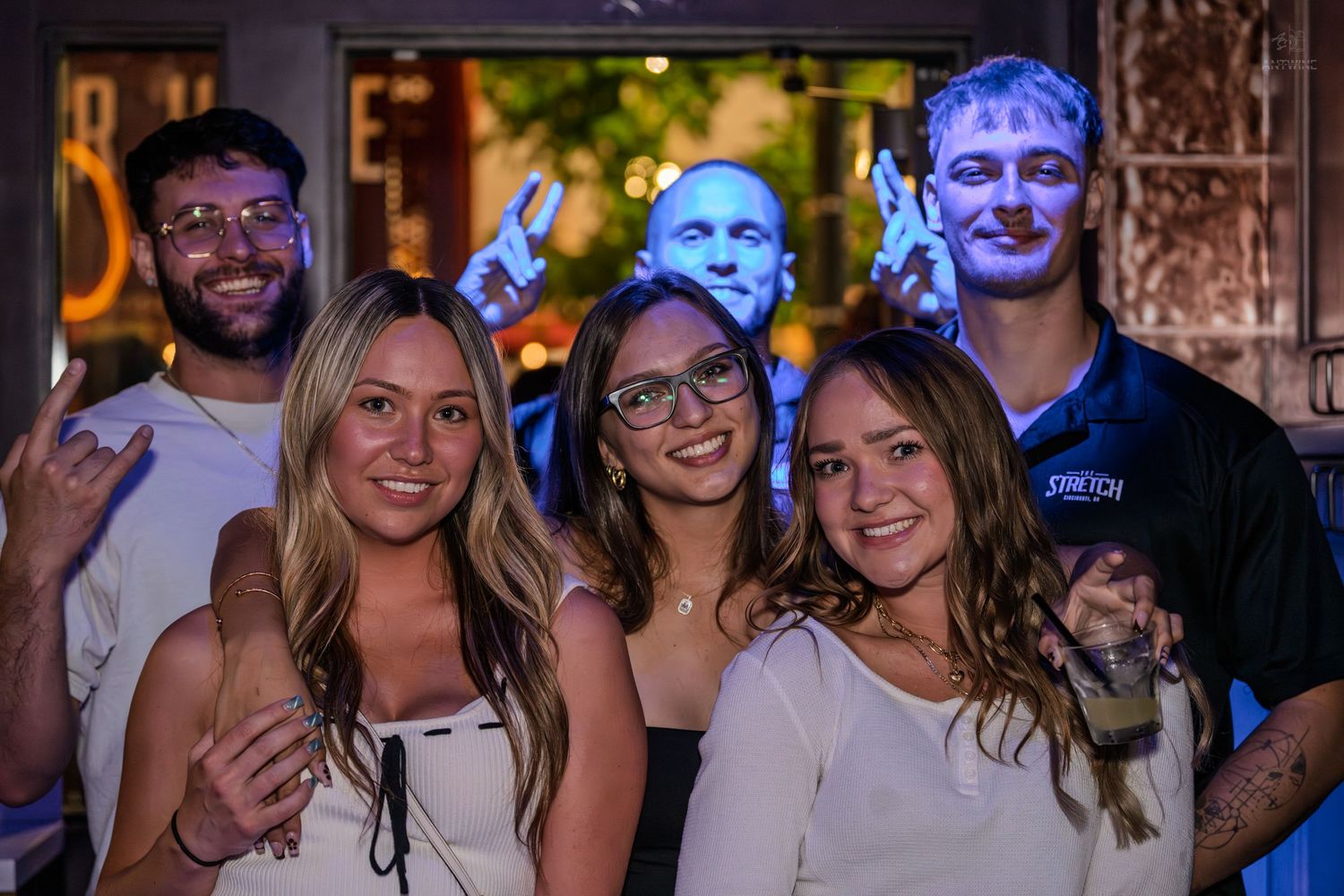 Group of six friends smiling and posing together outside a bar at night.