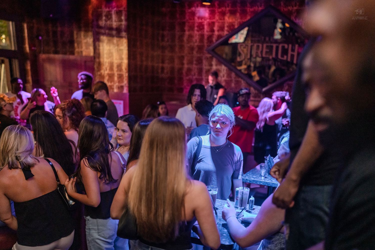 Nightclub crowd dancing under purple lights with a glowing bar area in the background.