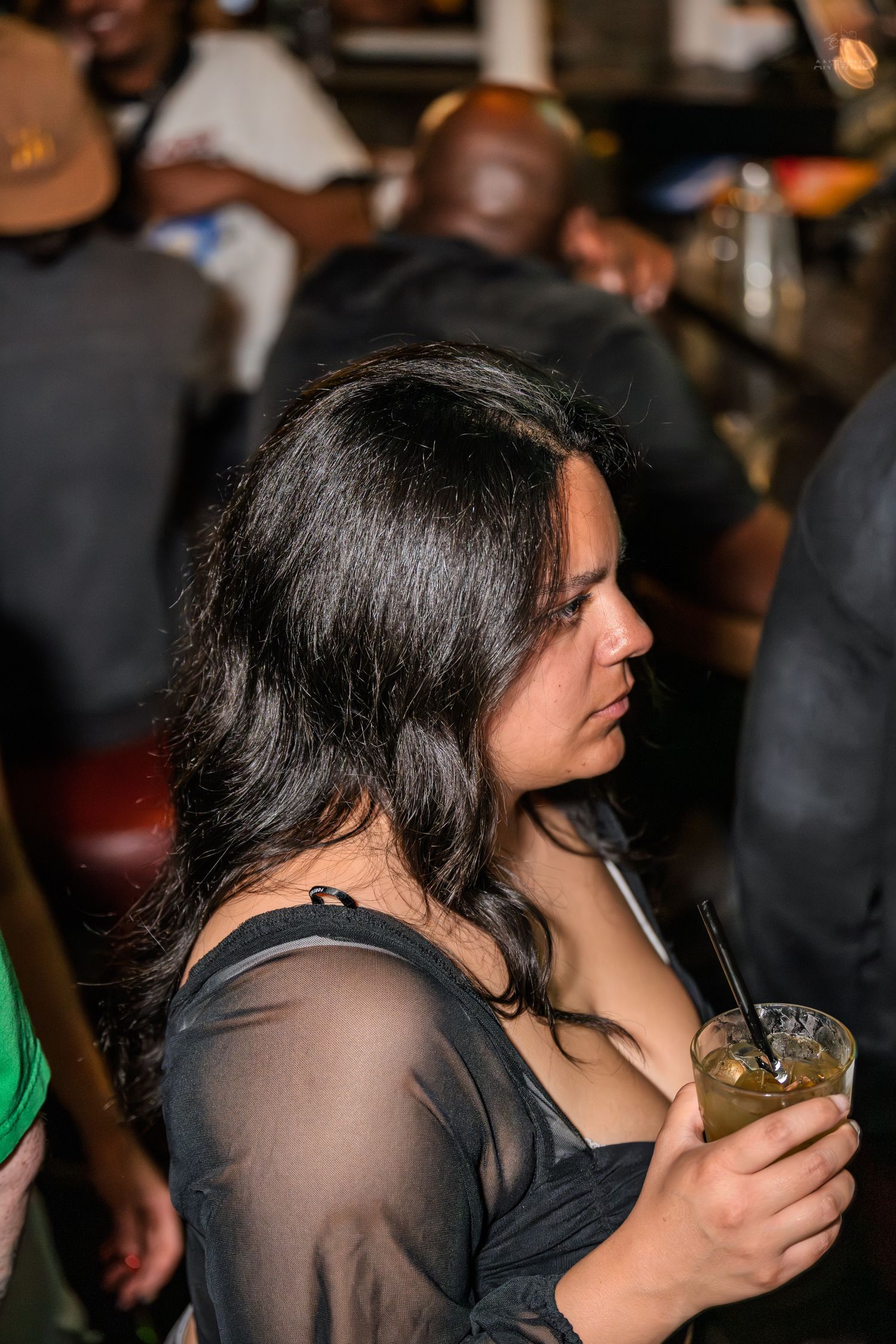 Woman in sheer black top holding a cocktail at a busy bar.