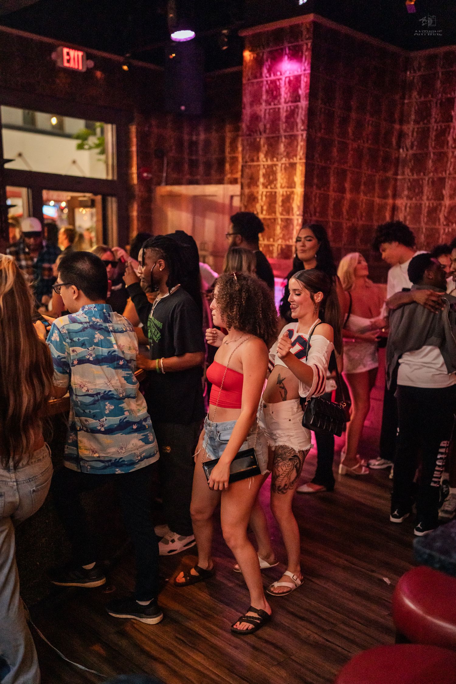 Diverse crowd socializing at a vibrant nightclub with pink neon lights and patterned walls.