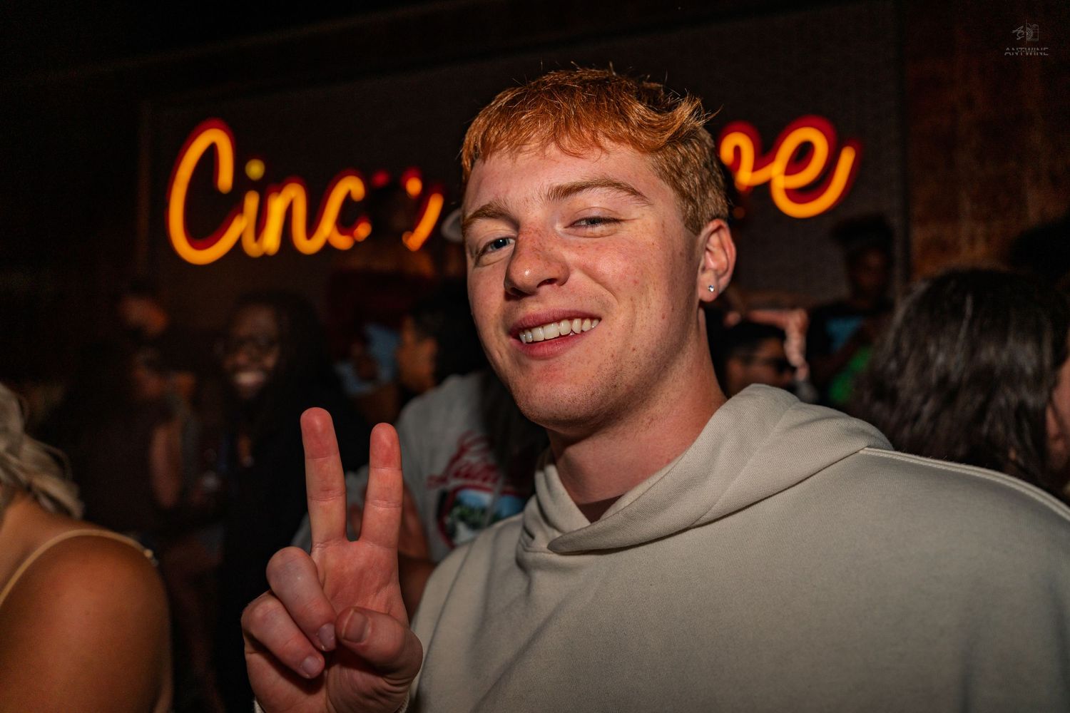 Cheerful young redhead man making a peace sign at a nightclub with neon signage behind him.