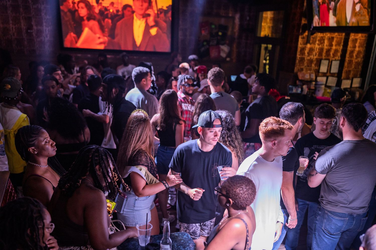 Crowded nightclub with diverse group of people socializing under warm lights with large screen projection.