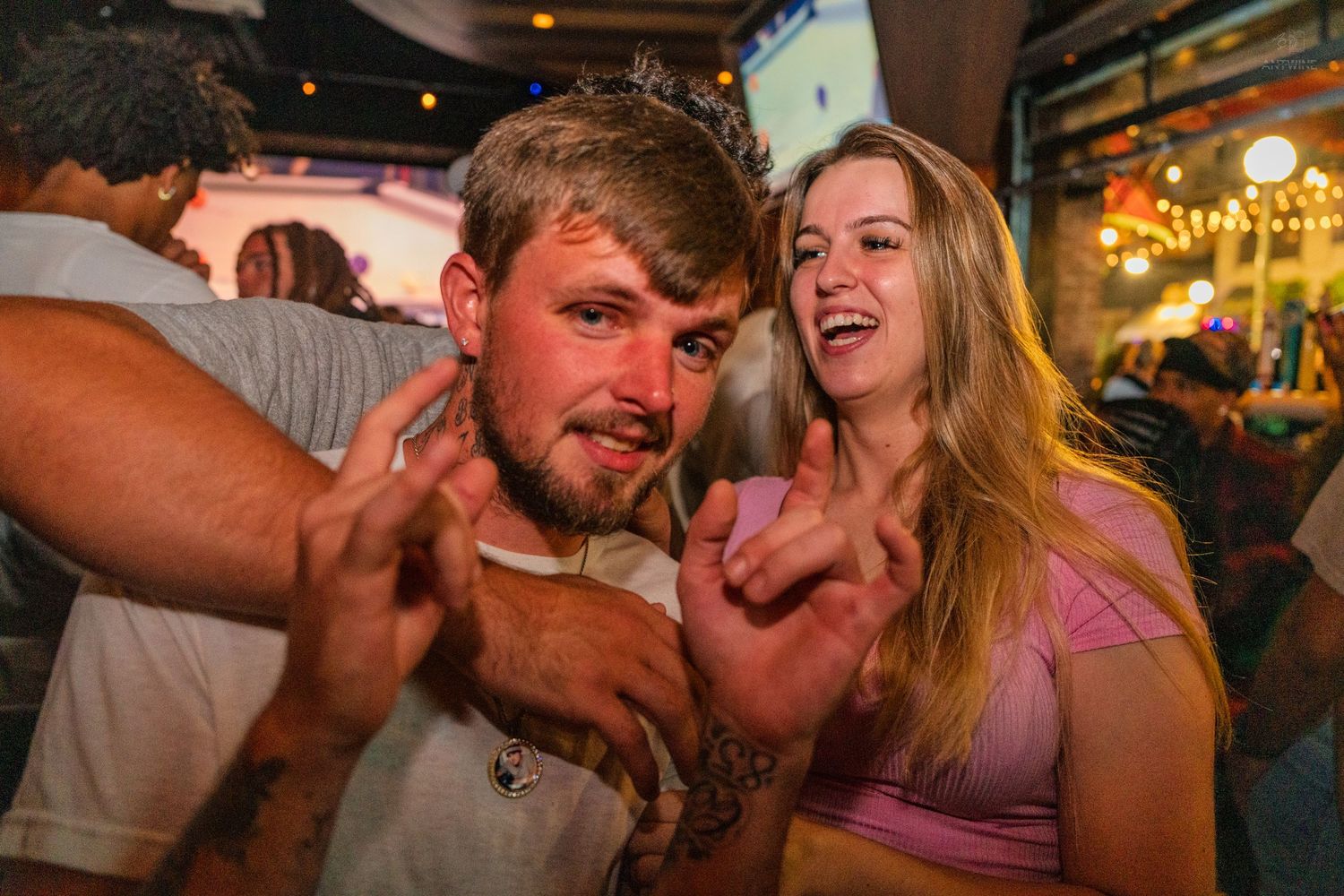 Man and woman laughing and posing together at a crowded bar making hand gestures.