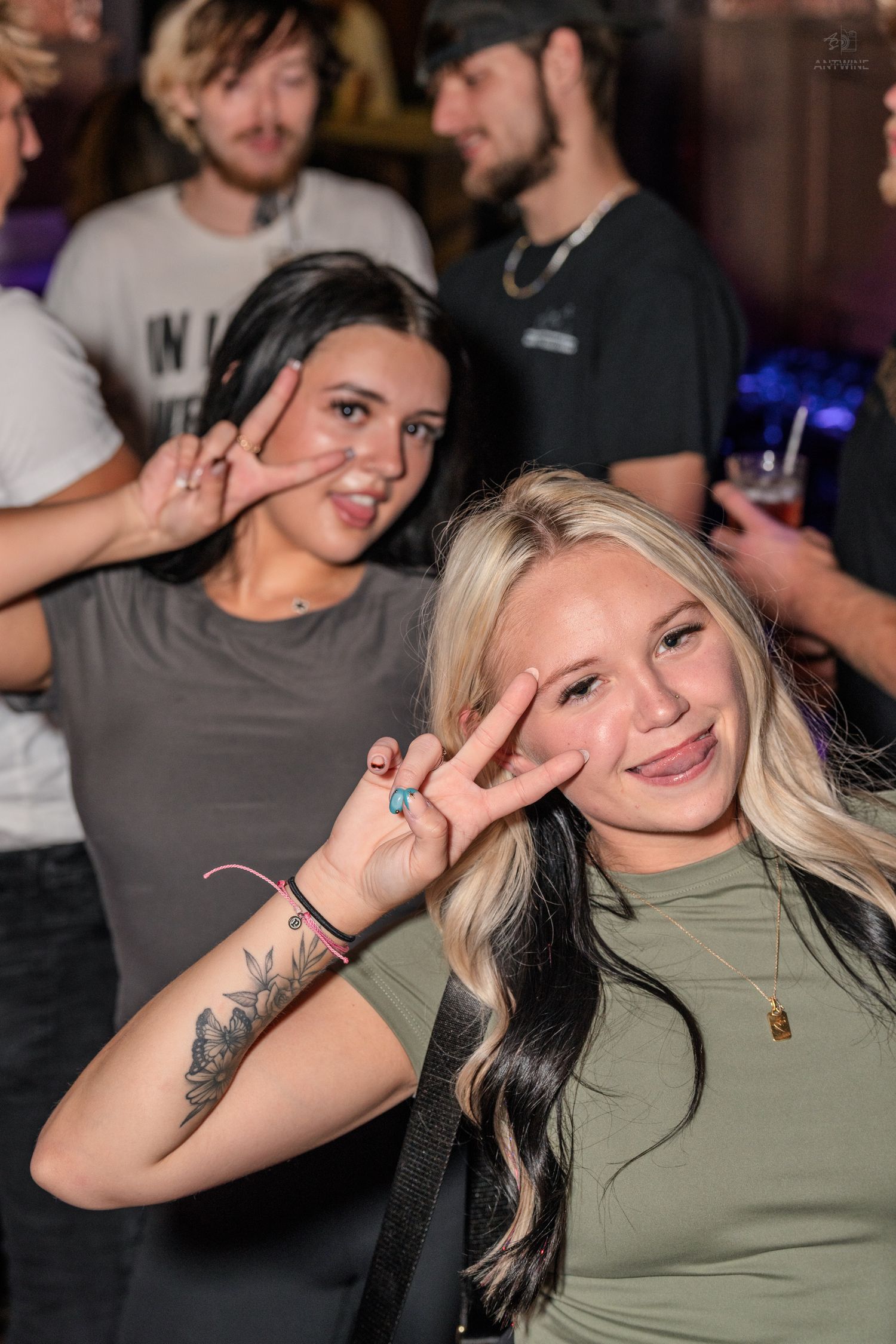Two young women posing with peace signs at a nightclub, one with dark hair and flower tattoo, one blonde.