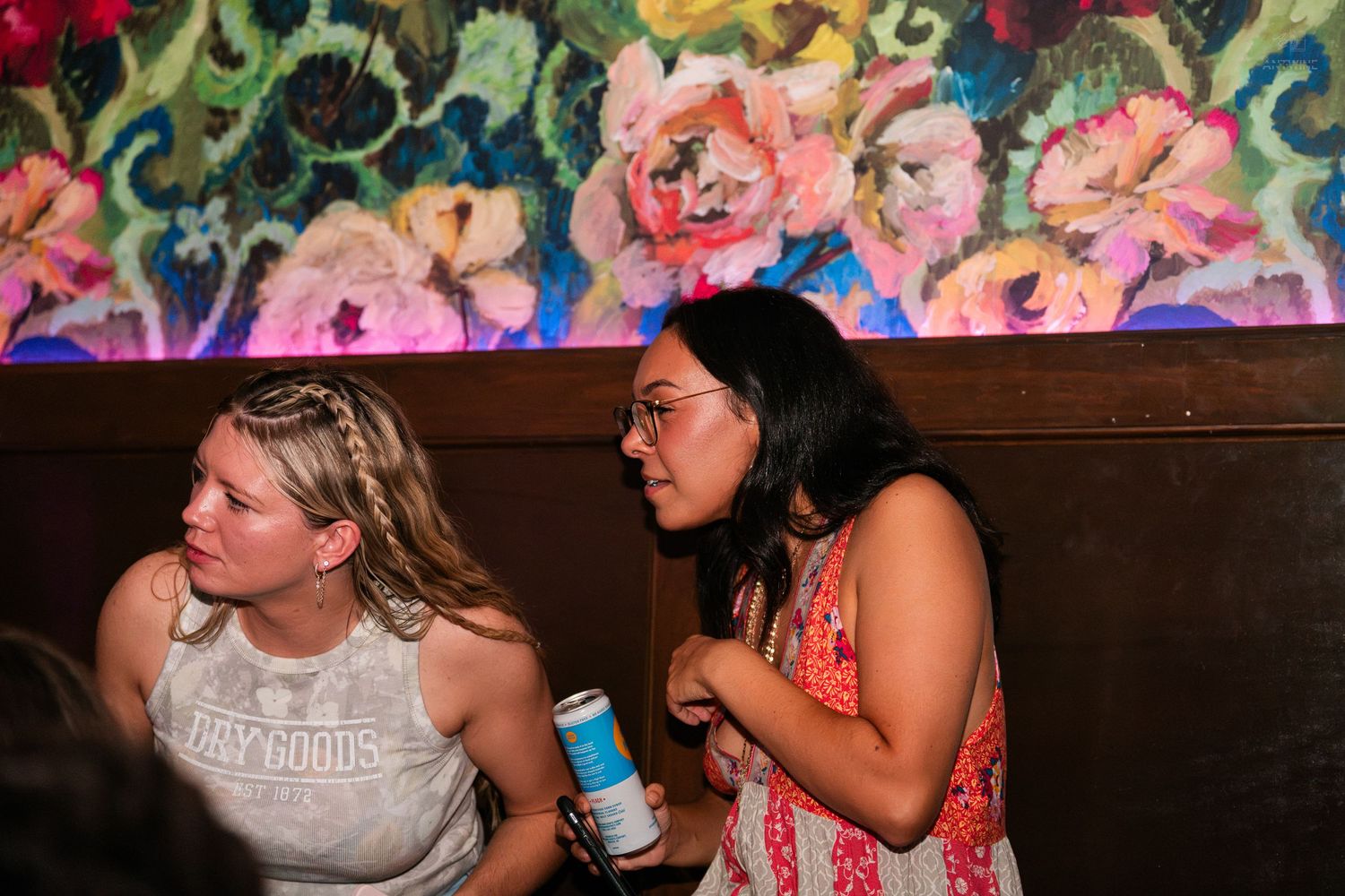 Two women conversing at a bar booth, one holding a can, with vibrant floral wall art behind them.