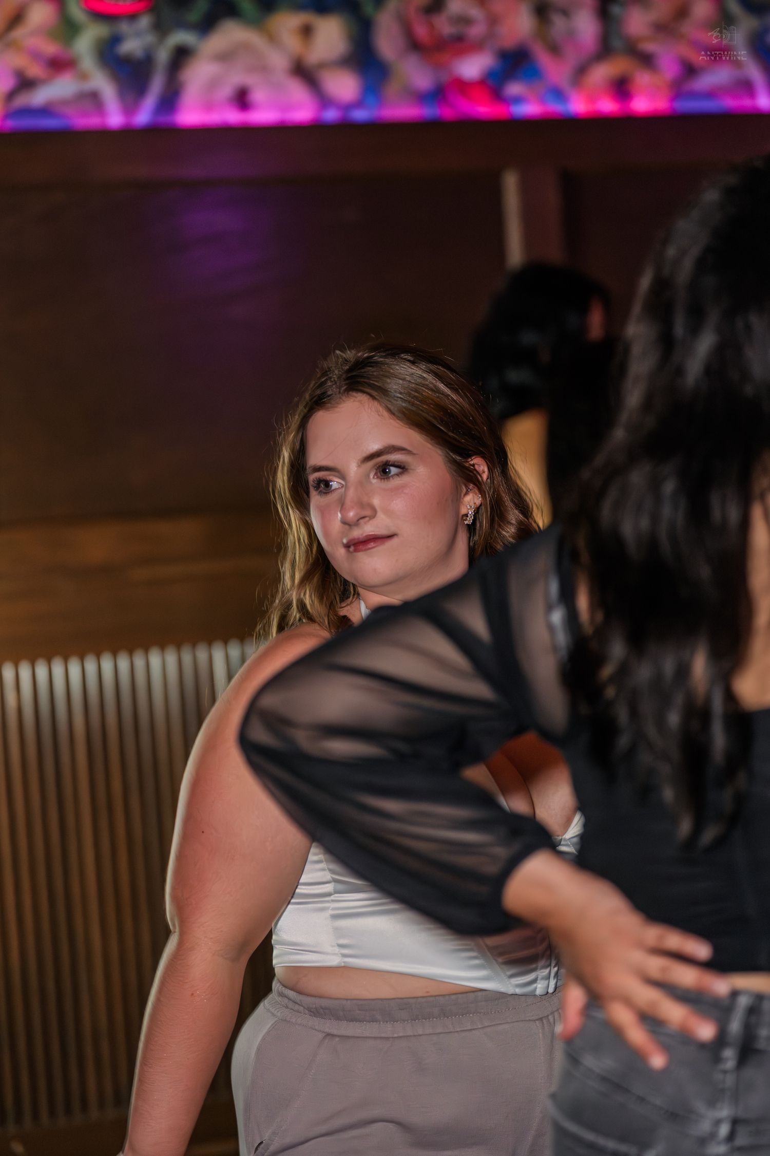 Young blonde woman in white crop top glances back at camera inside a dimly lit nightclub venue.
