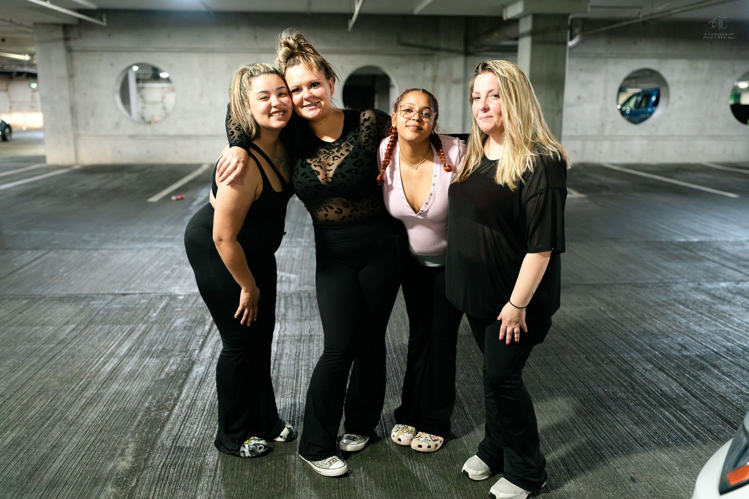 Four women embrace and smile together in a parking garage before a night out.