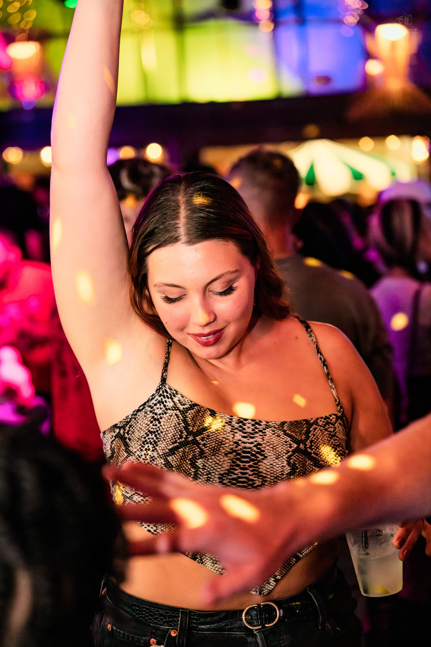 Woman in snakeskin crop top dancing with arm raised at a colorful nightclub.