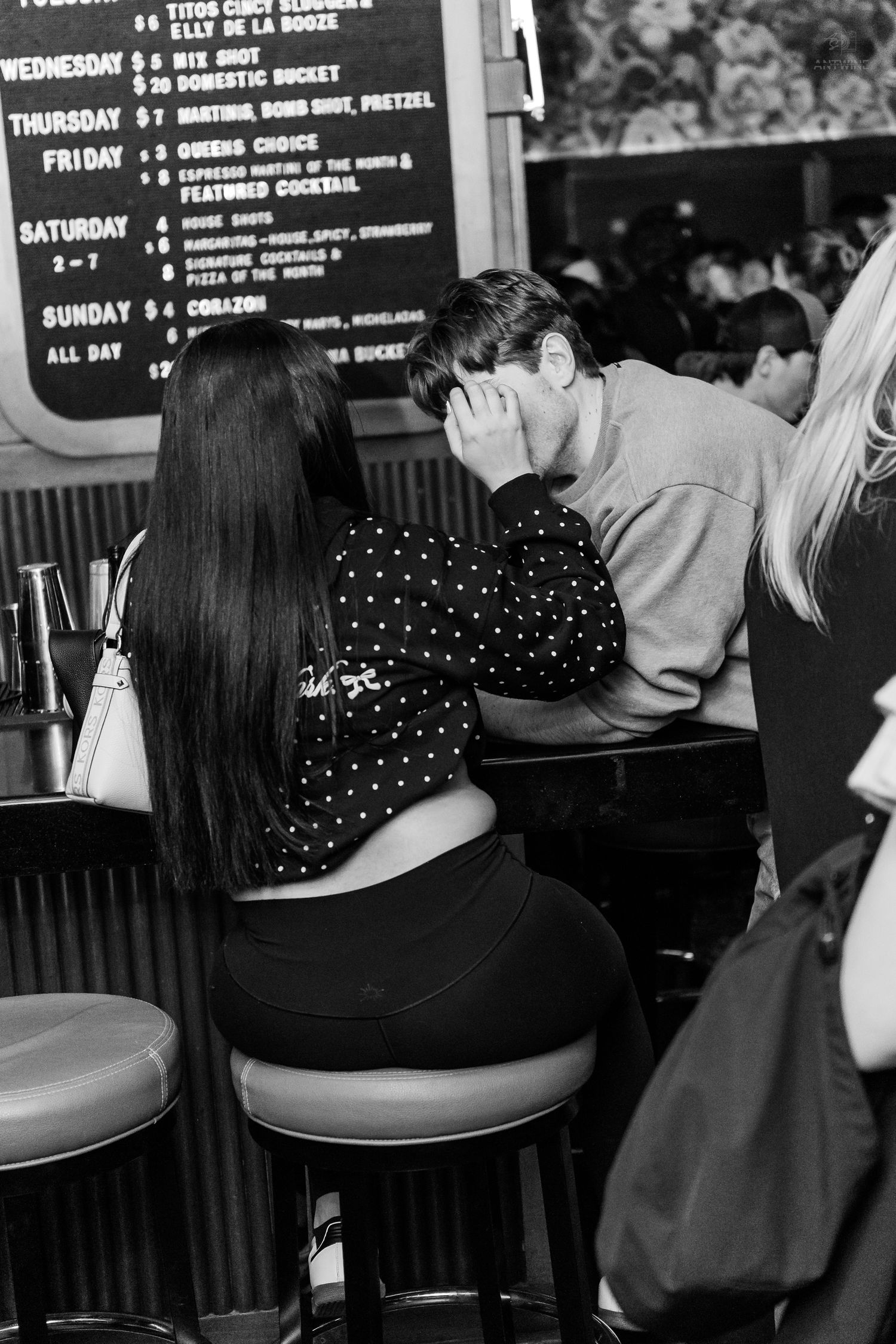 Couple sitting closely together at a bar counter with a drink specials chalkboard menu behind them.