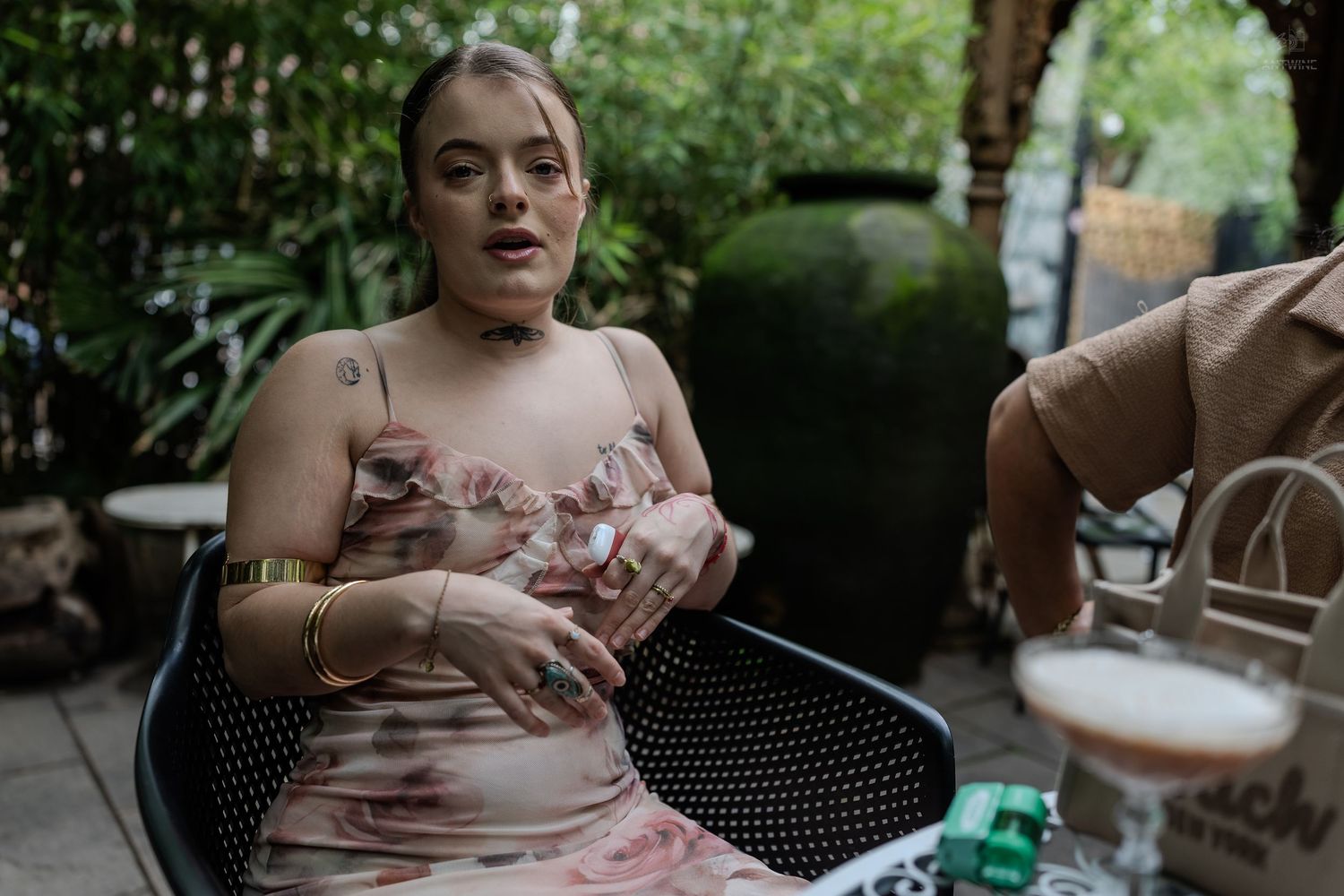 Woman in floral dress sitting outdoors at cafe table gesturing while talking.