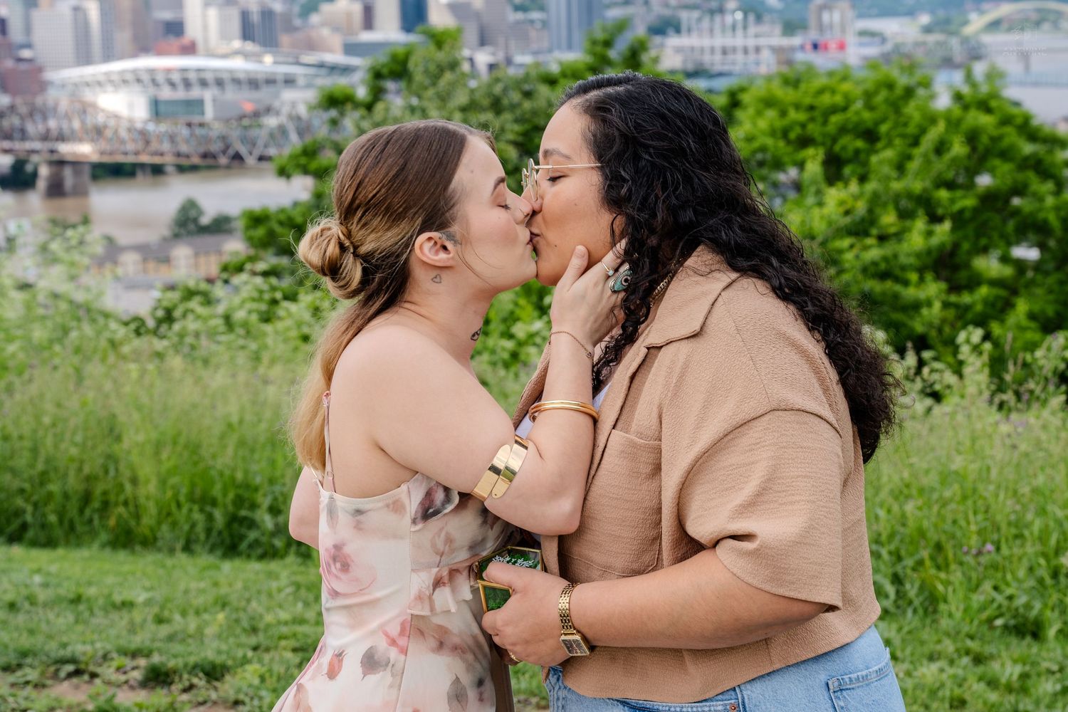 Two women sharing a romantic kiss outdoors with a lush green park and city bridge in the background.