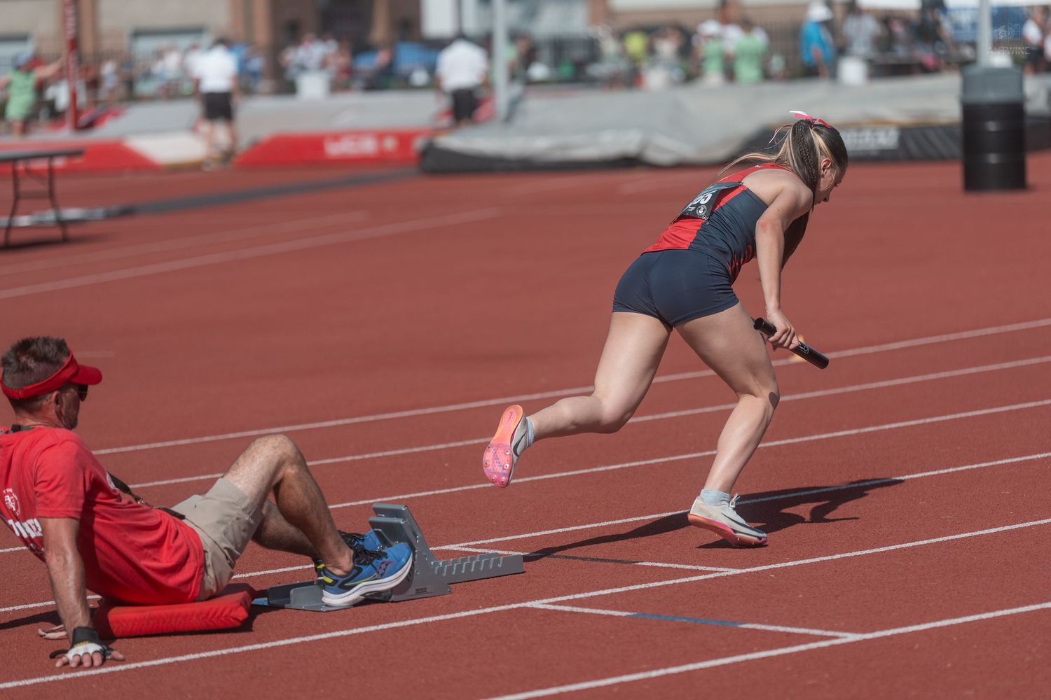 Female sprinter explodes from starting blocks on red track as coach observes during outdoor meet.
